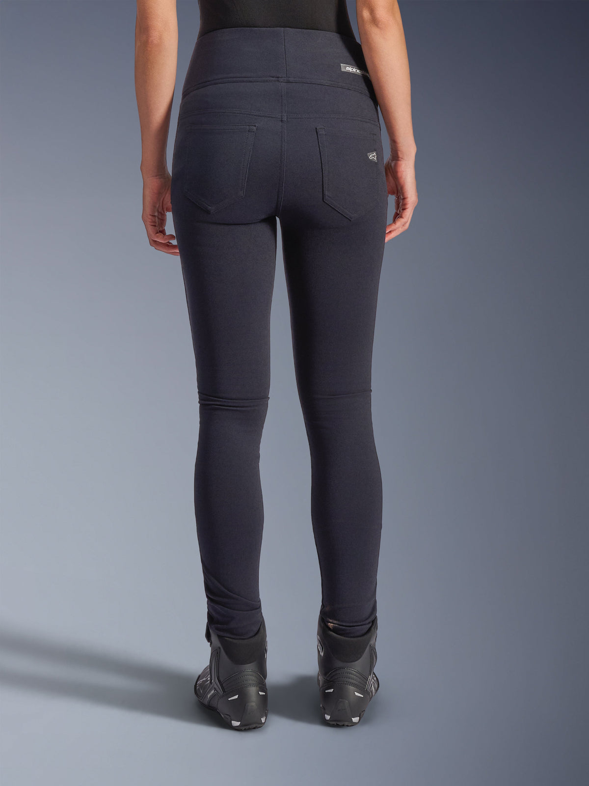 Alpinestars Stella Flex-AST Leggings, urbane Motorradhose, Schwarz, Rückansicht mit hoch geschnittener Skinny-Passform, zwei funktionelle Gesäßtaschen mit dezentem Branding, Stretch-Textilmaterial, kombiniert mit schwarzen Motorradschuhen.