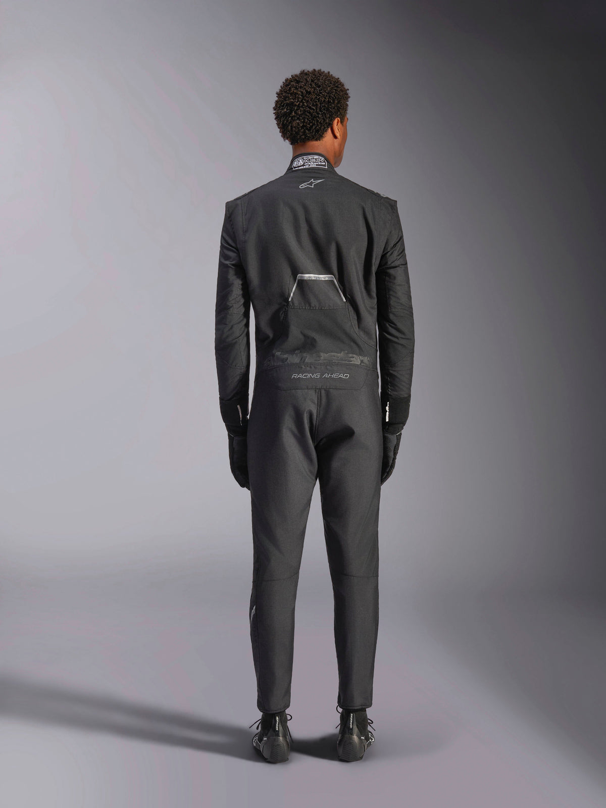 Alpinestars Hypertech V3 Anzug, Schwarz Dark Shadow, Rückansicht eines professionellen einteiligen Auto-Rennanzugs, mit FIA-Zertifizierungsetikett am Kragen und „Alpinestars“- sowie „RACING AHEAD“-Branding am unteren Rücken, getragen mit passenden schwarzen Rennhandschuhen und Stiefeln.