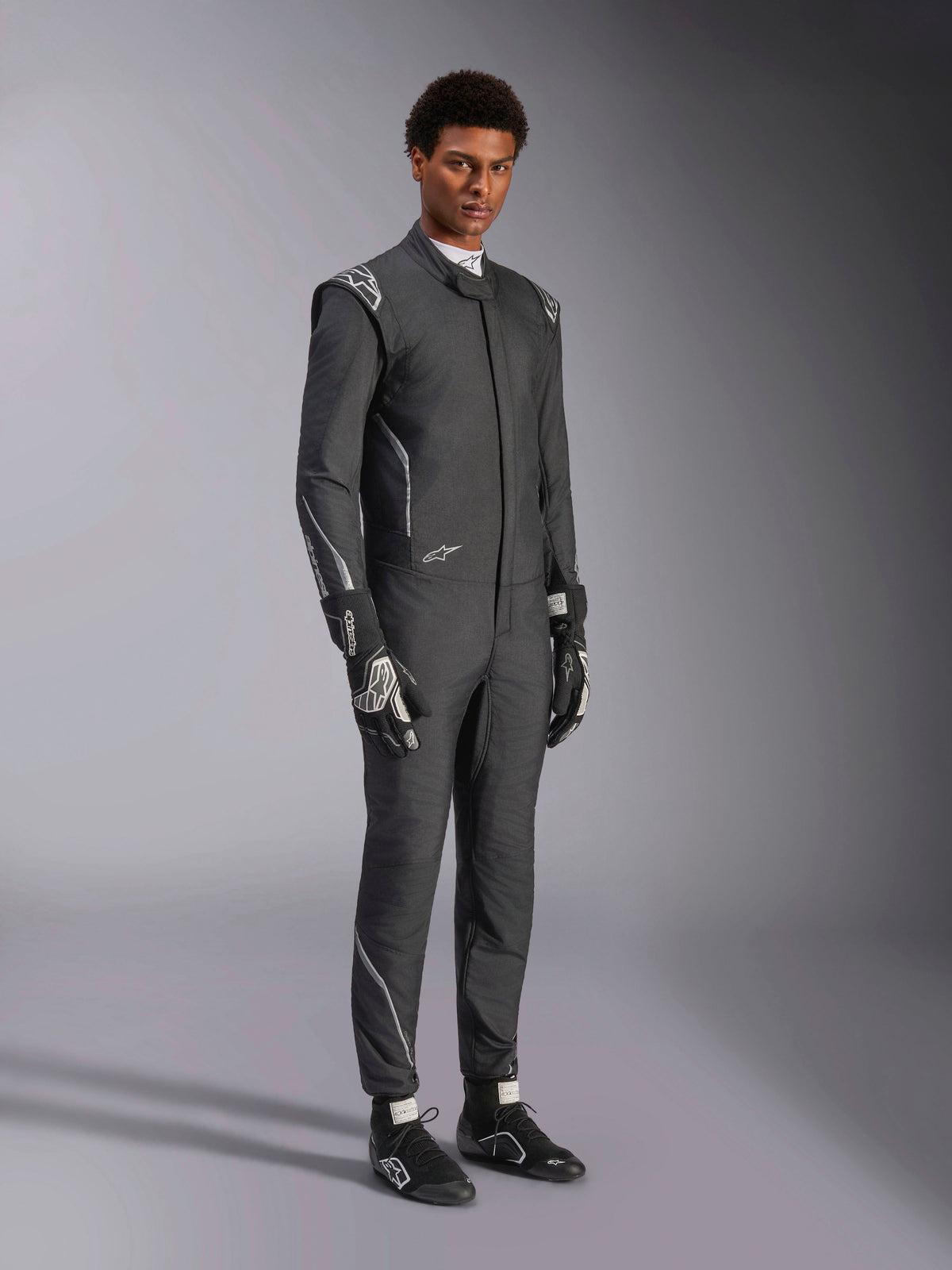 Alpinestars Hypertech V3 Anzug, Auto-Rennanzug, Schwarz und Dark Shadow, getragen von einem männlichen Model mit passenden schwarzen Handschuhen und Rennschuhen, sportlich schlanke Passform, mit Alpinestars-Logos und hellgrauen Akzenten, professionelle Motorsport-Schutzausrüstung