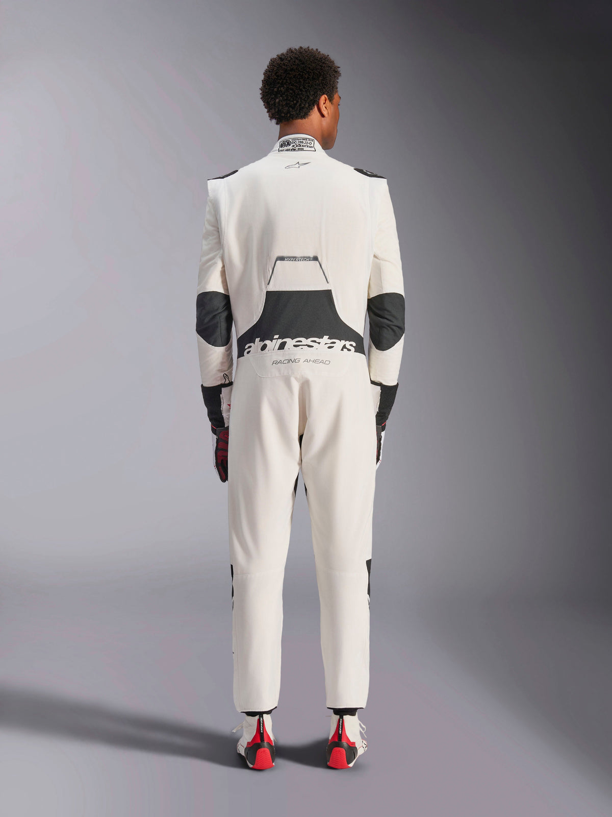 Rear view of a model wearing the Alpinestars Hypertech V3 Suit in White Black Silver, showcasing the large black lumbar stretch panel with 'alpinestars' and 'RACING AHEAD' branding, black elbow inserts, FIA certification label on the collar, and an athletic-slim fit designed for professional racing.