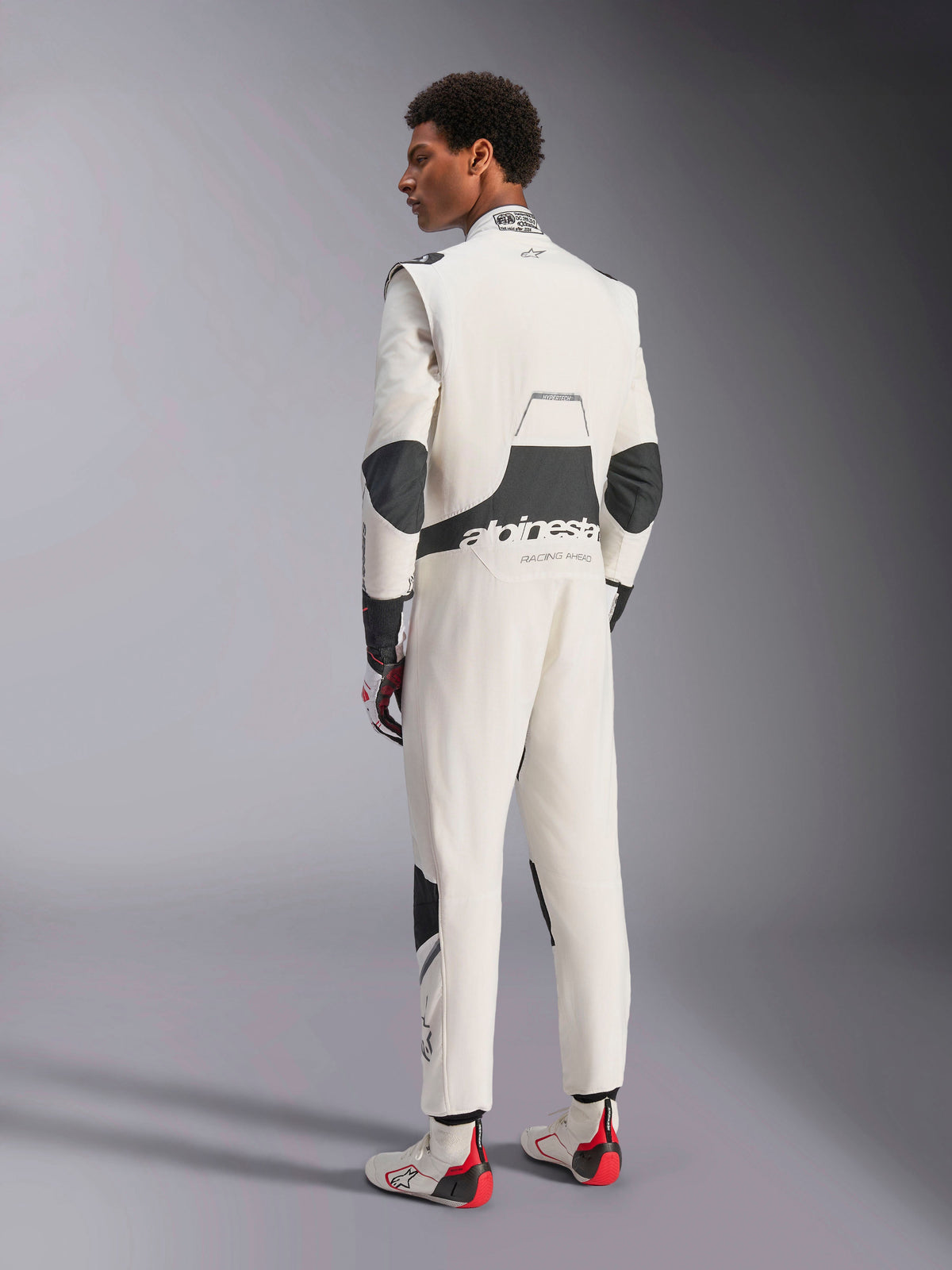 Alpinestars Hypertech V3 Suit, professional auto racing suit, White Black and Silver, rear view of model showing black stretch panels on the lumbar and elbows, Alpinestars branding on the back, paired with matching racing gloves and boots for a complete motorsport look