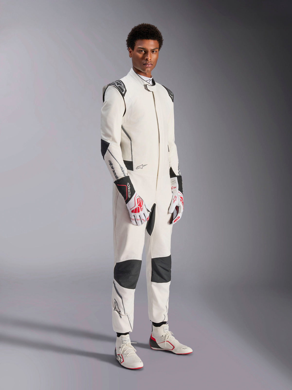 Male model wearing the Alpinestars Hypertech V3 Suit, White with Black and Silver accents, professional auto racing gear featuring black panels on shoulders and knees, styled with white and red racing gloves and white racing boots