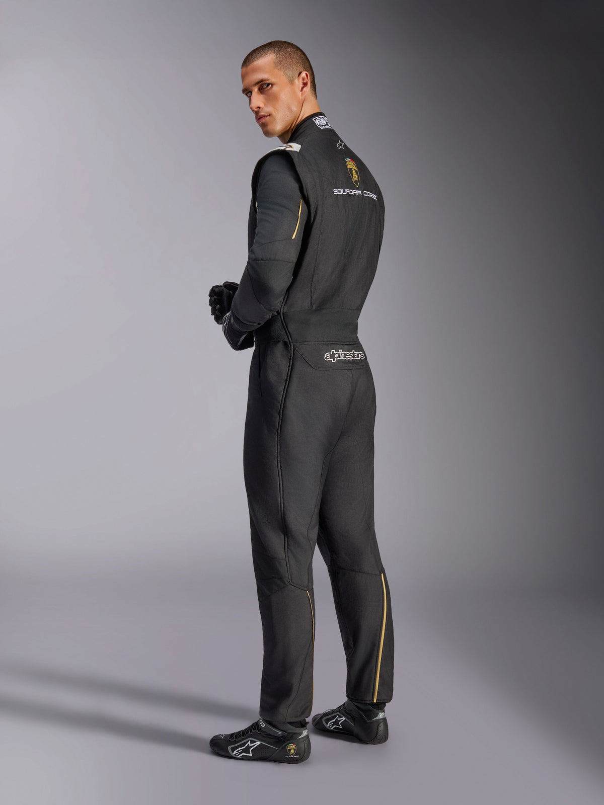 Alpinestars Limited Edition Lamborghini GP Tech V4 Anzug, professioneller Rennanzug, Schwarz mit silbernen und goldenen Akzenten, Rückansicht mit Lamborghini Squadra Corse und Alpinestars Branding, getragen mit schwarzen Rennhandschuhen und Rennschuhen, elegantes Motorsport-Design