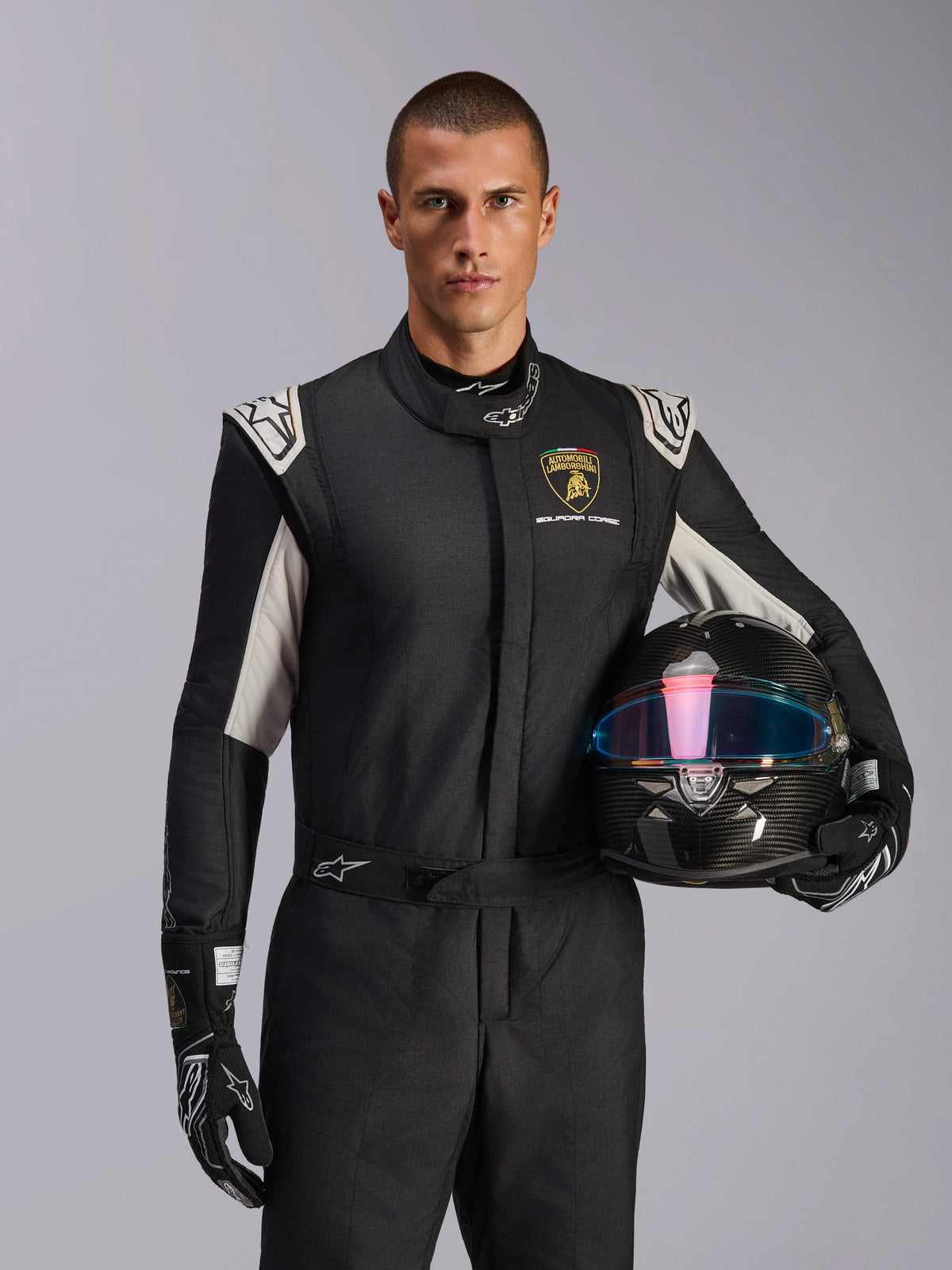 Alpinestars Limited Edition Lamborghini GP Tech V4 Anzug, Autorennanzug, Schwarz, Silber und Gold, getragen von einem Modell, das einen Carbon-Helm mit irisierendem Visier hält, mit Lamborghini Squadra Corse Branding und weißen Schulterakzenten