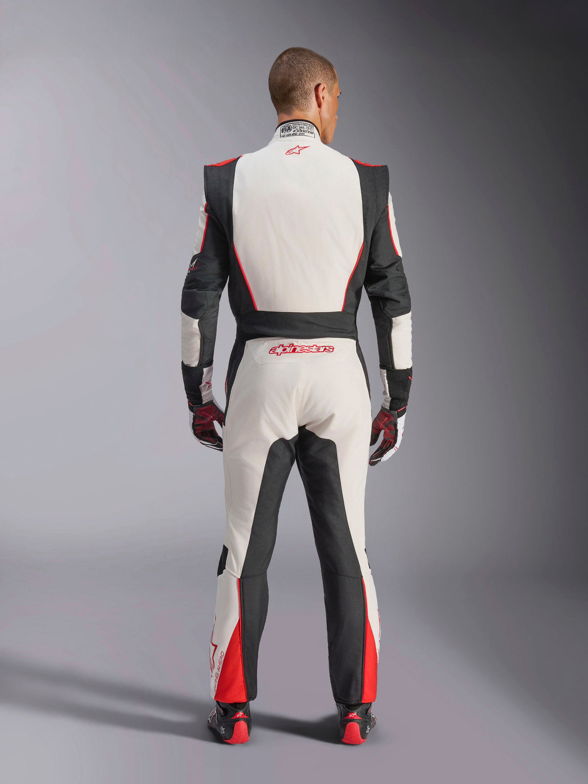 Alpinestars GP Tech V4 Anzug, Auto-Rennanzug, Weiß, Schwarz und Rot, Rückansicht eines Fahrers im einteiligen Anzug, mit schwarzen Stretcheinsätzen an den Seiten und Schultern, roten Akzenten an den Unterschenkeln, Alpinestars-Branding am unteren Rücken und am Kragen, professionelle FIA-zertifizierte Motorsportausrüstung