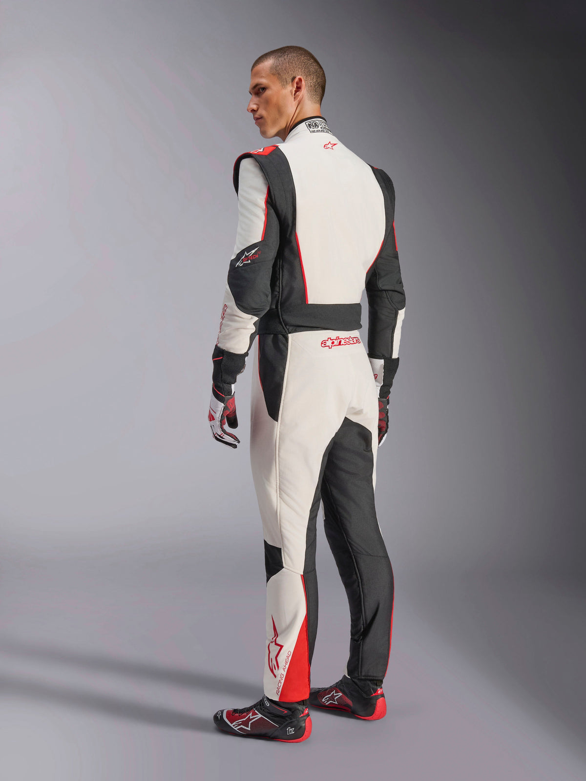 Alpinestars GP Tech V4 Anzug, Rennanzug in Weiß Schwarz Rot, Rückansicht mit ergonomischen Stretcheinsätzen am unteren Rücken und an den Beinen, kombiniert mit passenden Rennhandschuhen und -schuhen, professionelle Motorsportbekleidung.