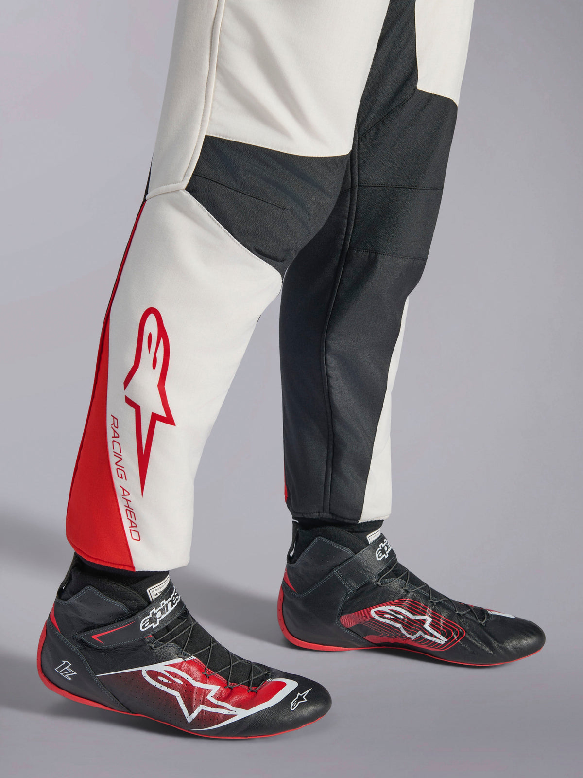 Alpinestars GP Tech V4 Anzug, Auto-Rennanzug, Weiß, Schwarz und Rot, Detailansicht der Unterschenkel mit weißen und schwarzen Paneelen, roten Akzenten und „RACING AHEAD“-Branding, getragen mit schwarzen Rennschuhen, professionelle Motorsportausrüstung