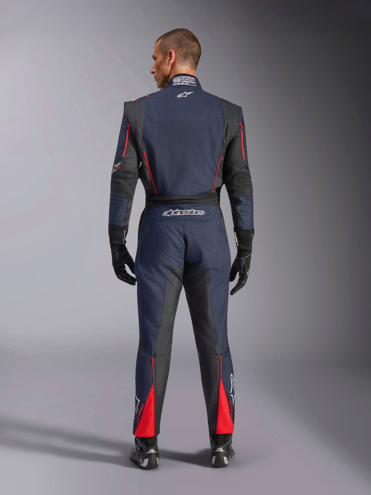 Rückansicht des Alpinestars GP Tech V4 Anzugs in Blue Navy Black Red, einteiliger Auto-Rennanzug mit schwarzen Stretcheinsätzen und roten Akzenten, mit Alpinestars-Branding und FIA-Zertifizierungsdetails, getragen mit professionellen Rennhandschuhen und Stiefeln.