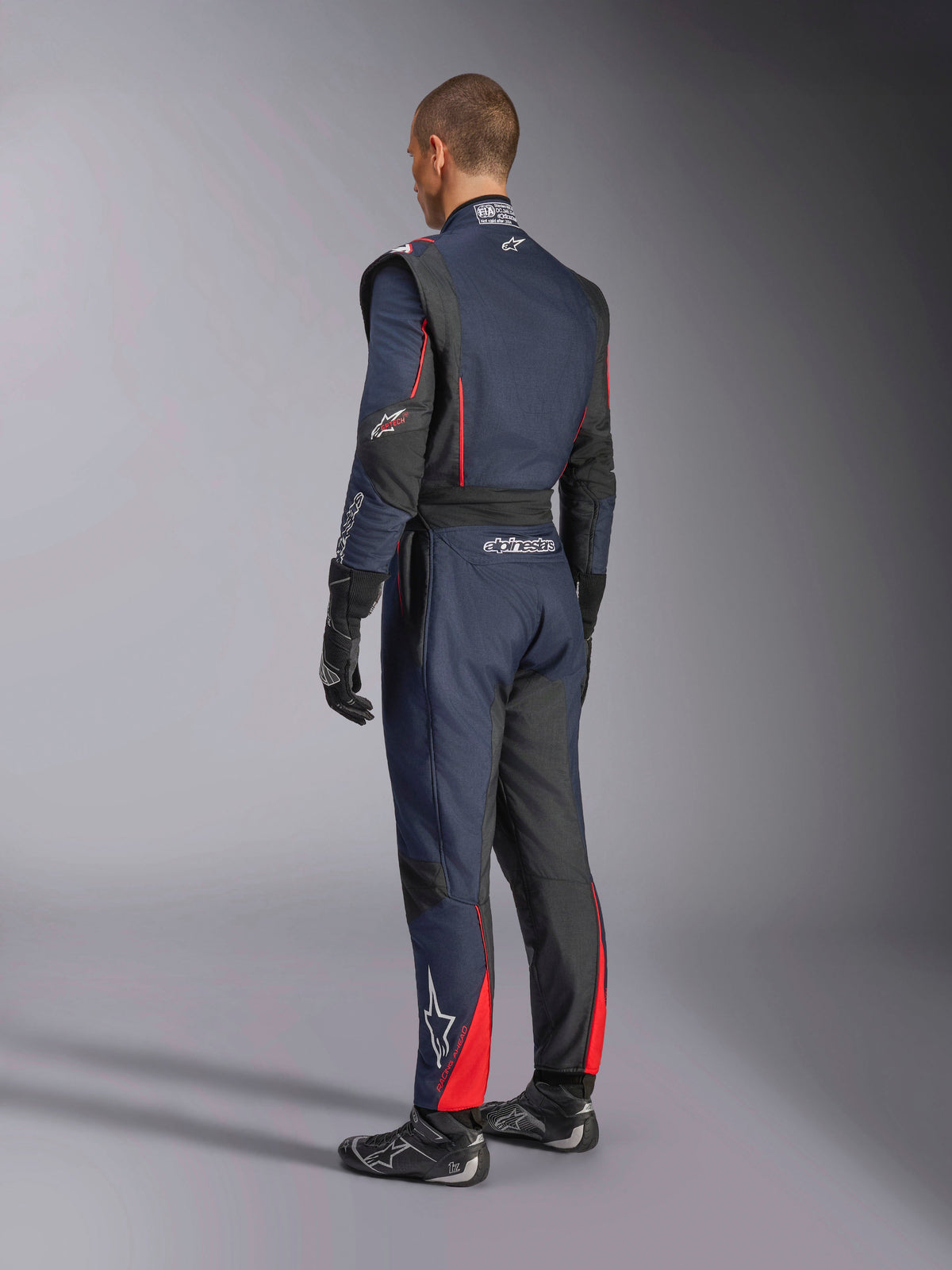 Alpinestars GP Tech V4 Anzug, Navy-Blau, Schwarz und Rot, Rückansicht eines professionellen einteiligen Autorennanzugs, mit roten Akzenten und schwarzen Einsätzen, getragen mit schwarzen Rennhandschuhen und Rennschuhen, FIA-zertifizierte Motorsportausrüstung.
