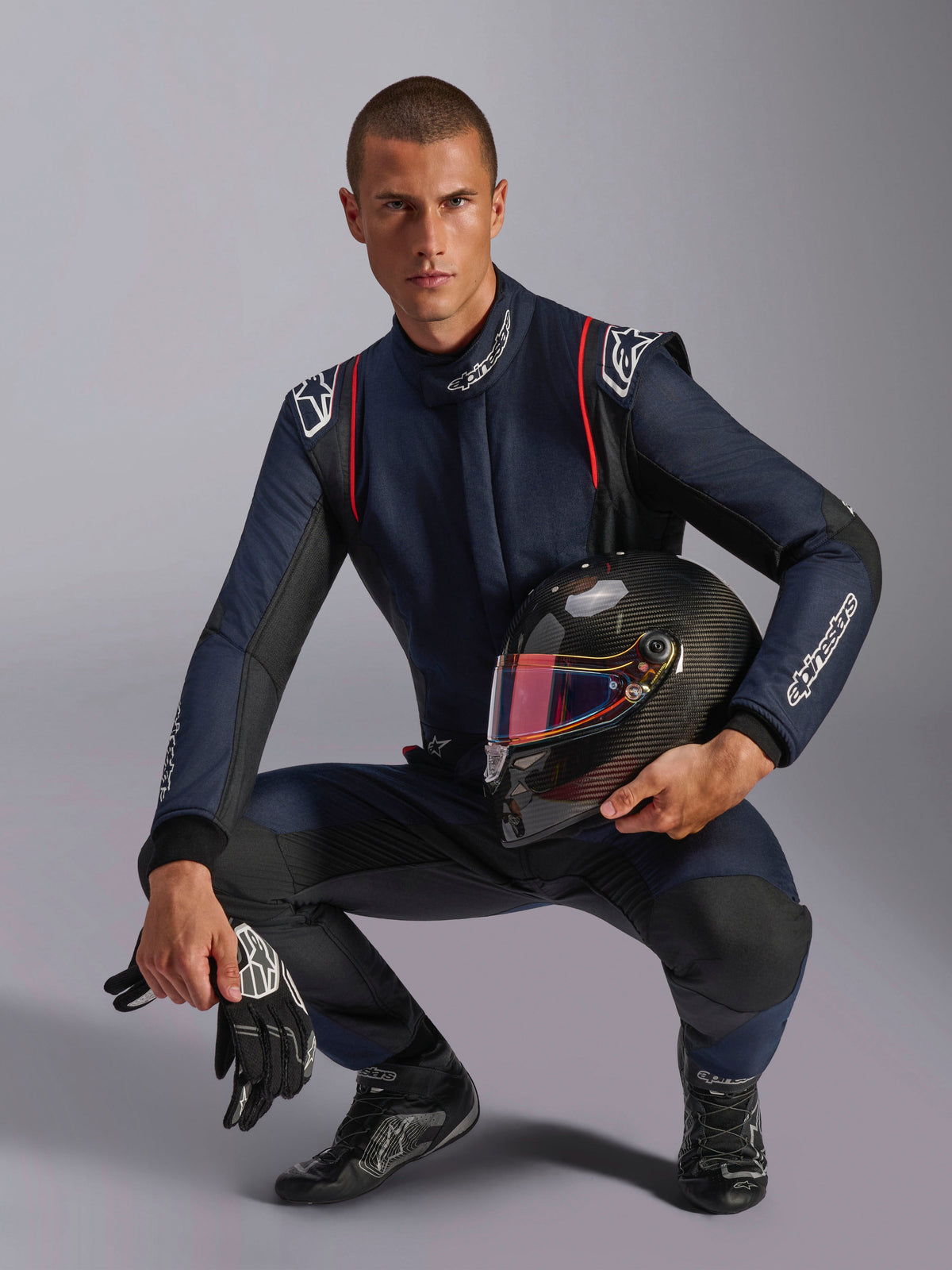 Männlicher Rennfahrer im Alpinestars GP Tech V4 Anzug, Marineblau mit schwarzen und roten Akzenten, hält einen Carbonhelm mit irisierendem Visier, schwarze Rennhandschuhe und Stiefel, professionelle Automobil-Rennausrüstung