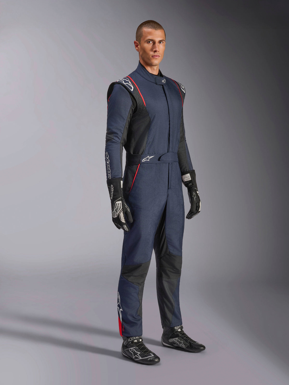 Alpinestars GP Tech V4 Anzug, einteiliger Auto-Rennanzug, Navy-Blau, Schwarz und Rot, getragen von einem Modell mit schwarzen Rennsport-Handschuhen und Stiefeln, mit schwarzen Einsätzen und roten Paspeln, professionelle Motorsportausrüstung.