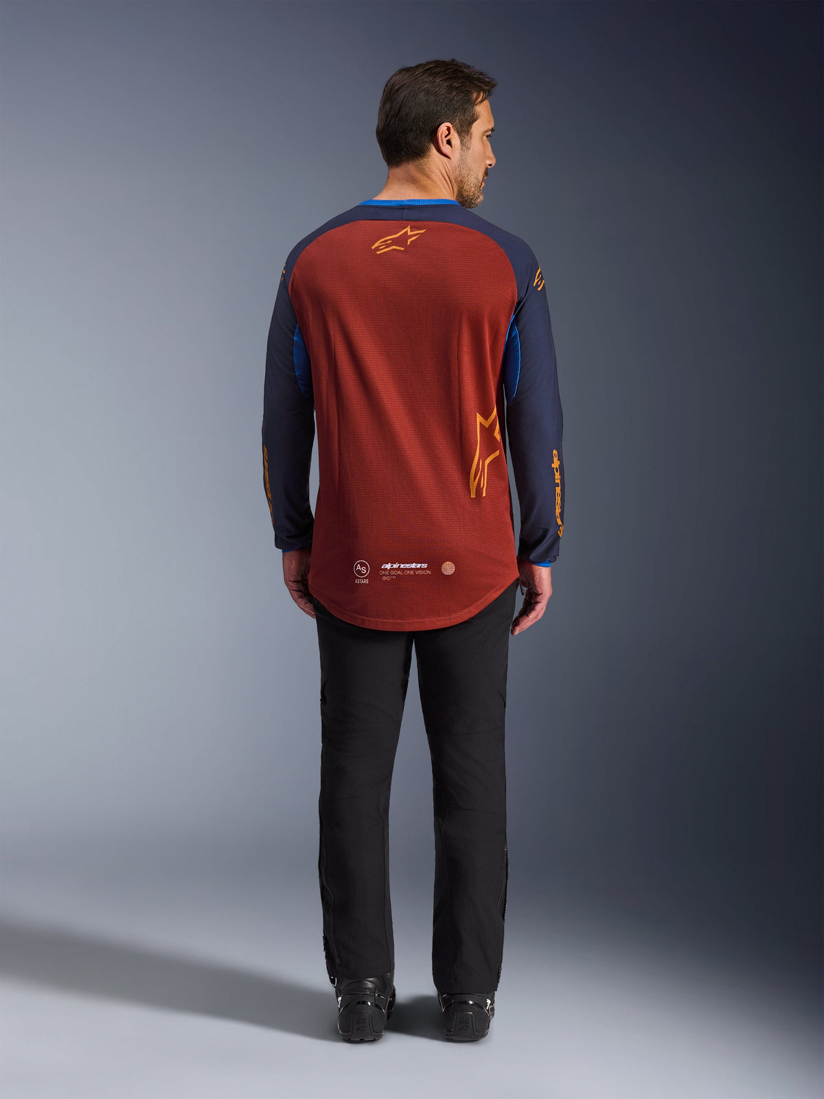 Alpinestars Maxdura Dual Jersey in Rust Brown und Mood Indigo, Rückansicht mit abgesenktem Saum im Rückenbereich und Logo-Details, getragen mit einer schwarzen technischen Hose und Stiefeln, konzipiert für Adventure- und Dual-Sport-Motorradfahren.