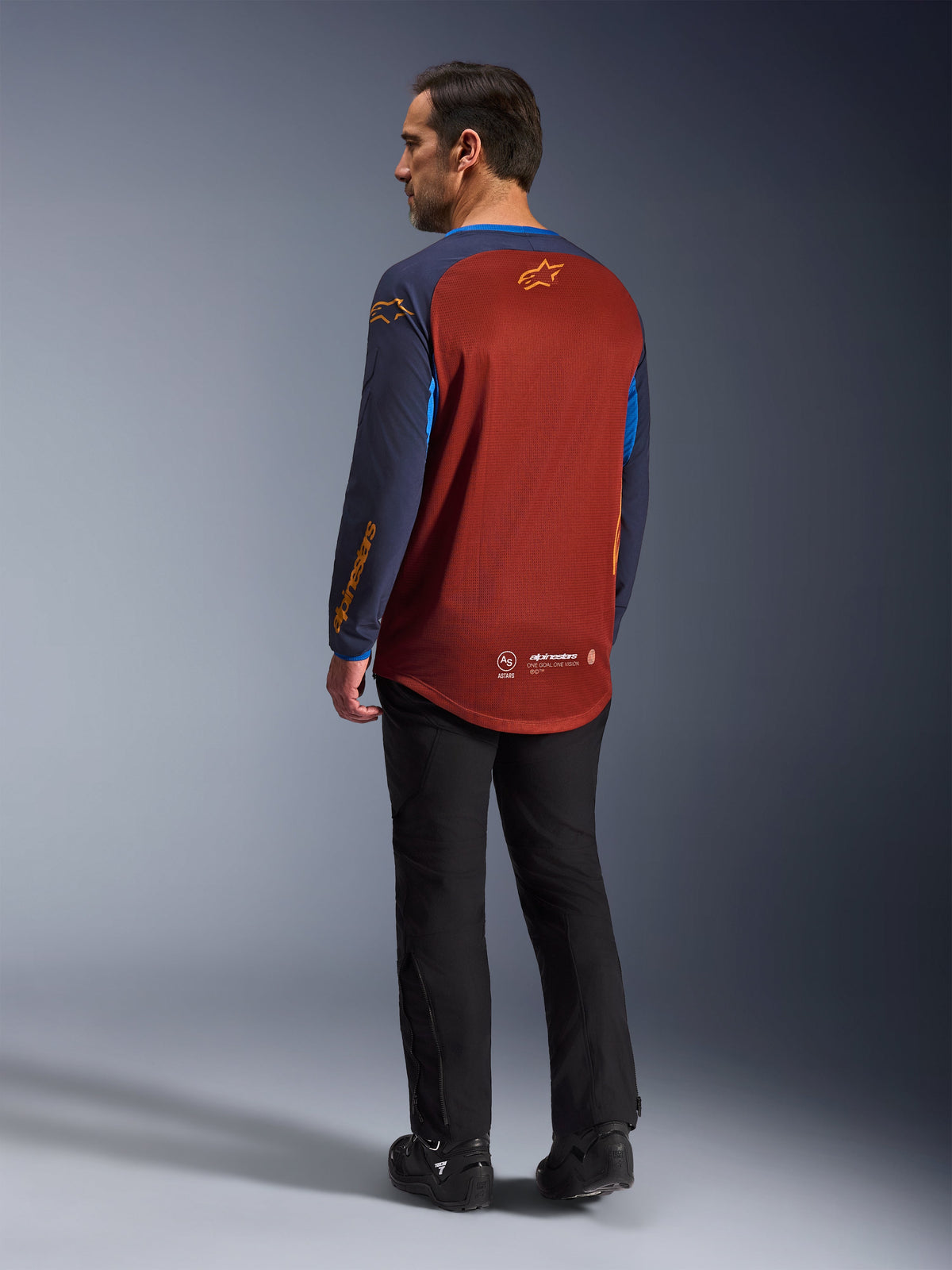Alpinestars Maxdura Dual Jersey, Rust Brown und Mood Indigo, Adventure-Motorrad-Jersey in der Rückansicht, rostbrauner Mesh-Einsatz zur Belüftung, navyblaue Ärmel mit orangefarbenen Logos, kombiniert mit schwarzer Motorradhose und Stiefeln, technische Offroad-Ausrüstung