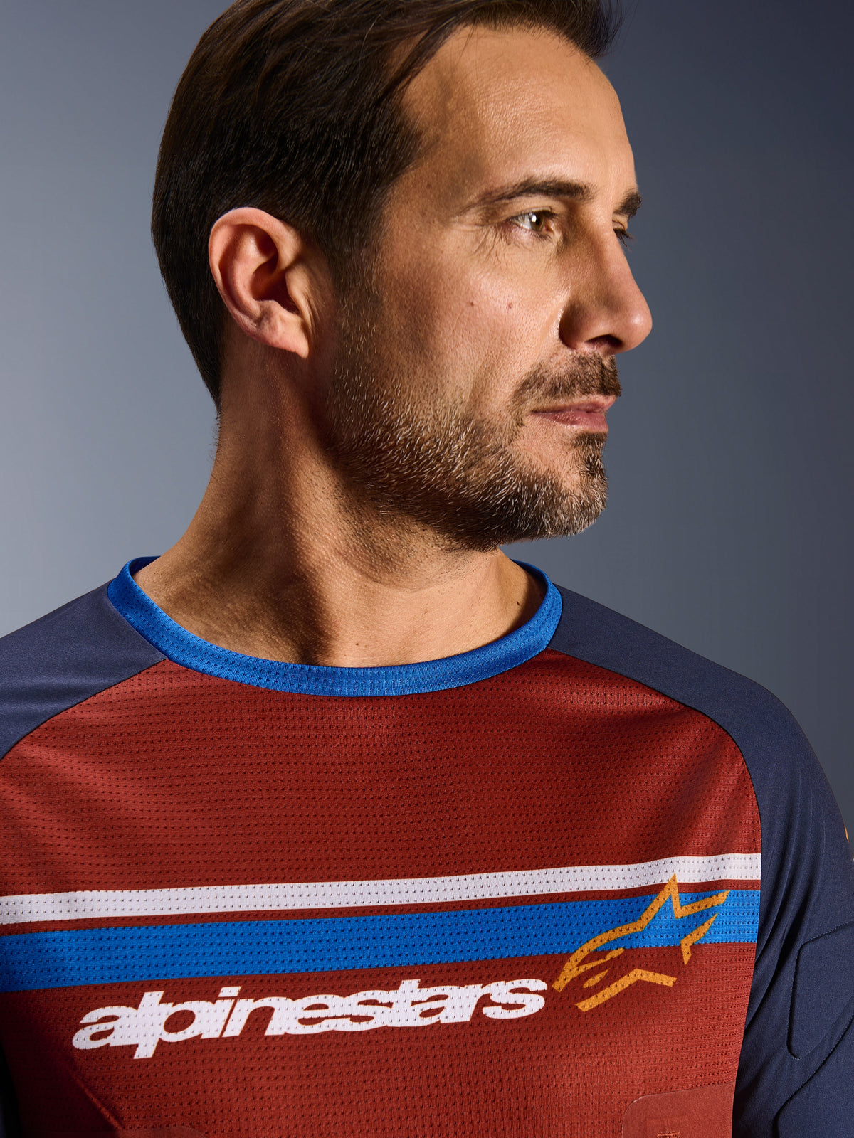 Alpinestars Maxdura Dual Trikot, Rostbraun und Mood Indigo, Nahaufnahme eines Fahrers, der das technische Mesh-Trikot trägt, mit horizontalen weißen und blauen Bruststreifen, weißem Alpinestars-Schriftzug und einem orangefarbenen Stern-Logo.