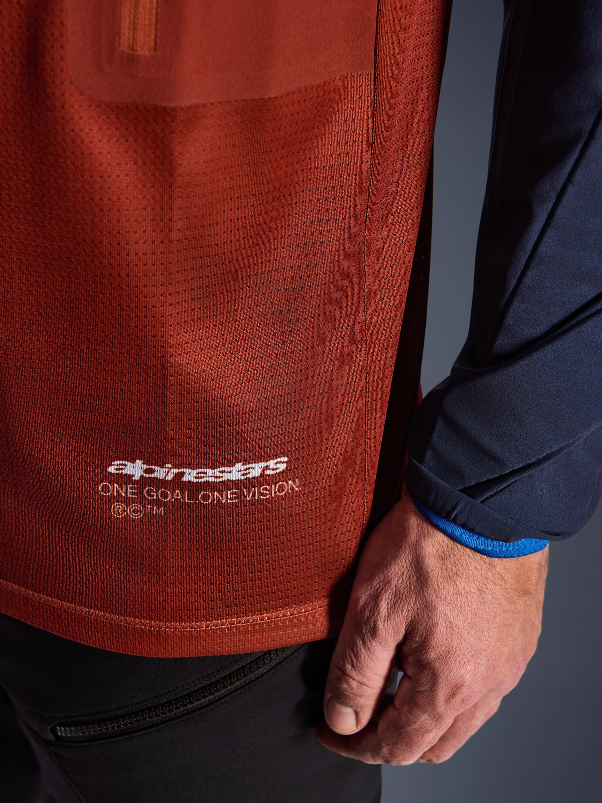 Alpinestars Maxdura Dual Trikot, Adventure-Motorradtrikot, Rostbraun und Mood Indigo, Nahaufnahme des Mesh-Torso-Gewebes und des navyfarbenen gewebten Ärmels, weißes Alpinestars „One Goal One Vision“-Branding am unteren Saum.