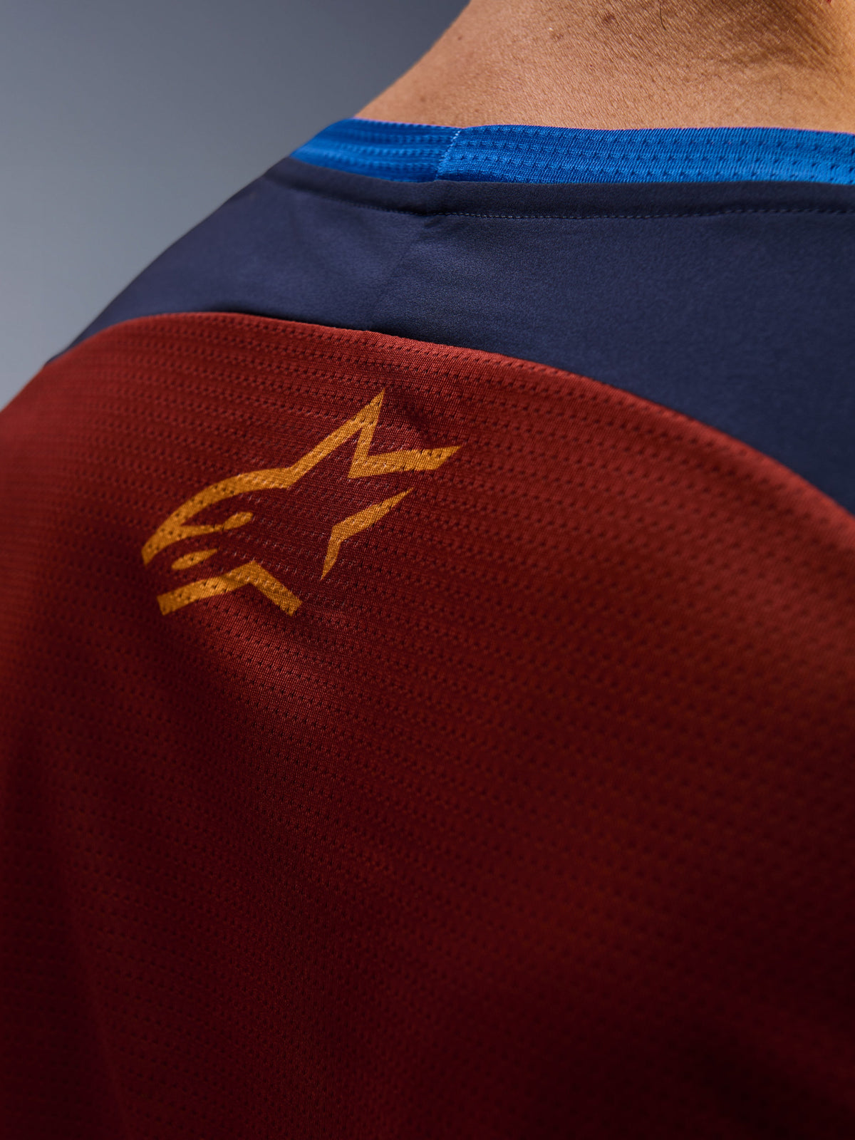 Nahaufnahme des Alpinestars Maxdura Dual Trikots, rostbraunes Mesh-Gewebe auf der Rückseite, kleines orangefarbenes Stern-Logo, dunkelblauer oberer Einsatz und hellblauer Kragenbesatz, Detailansicht eines Adventure-Motorradtrikots