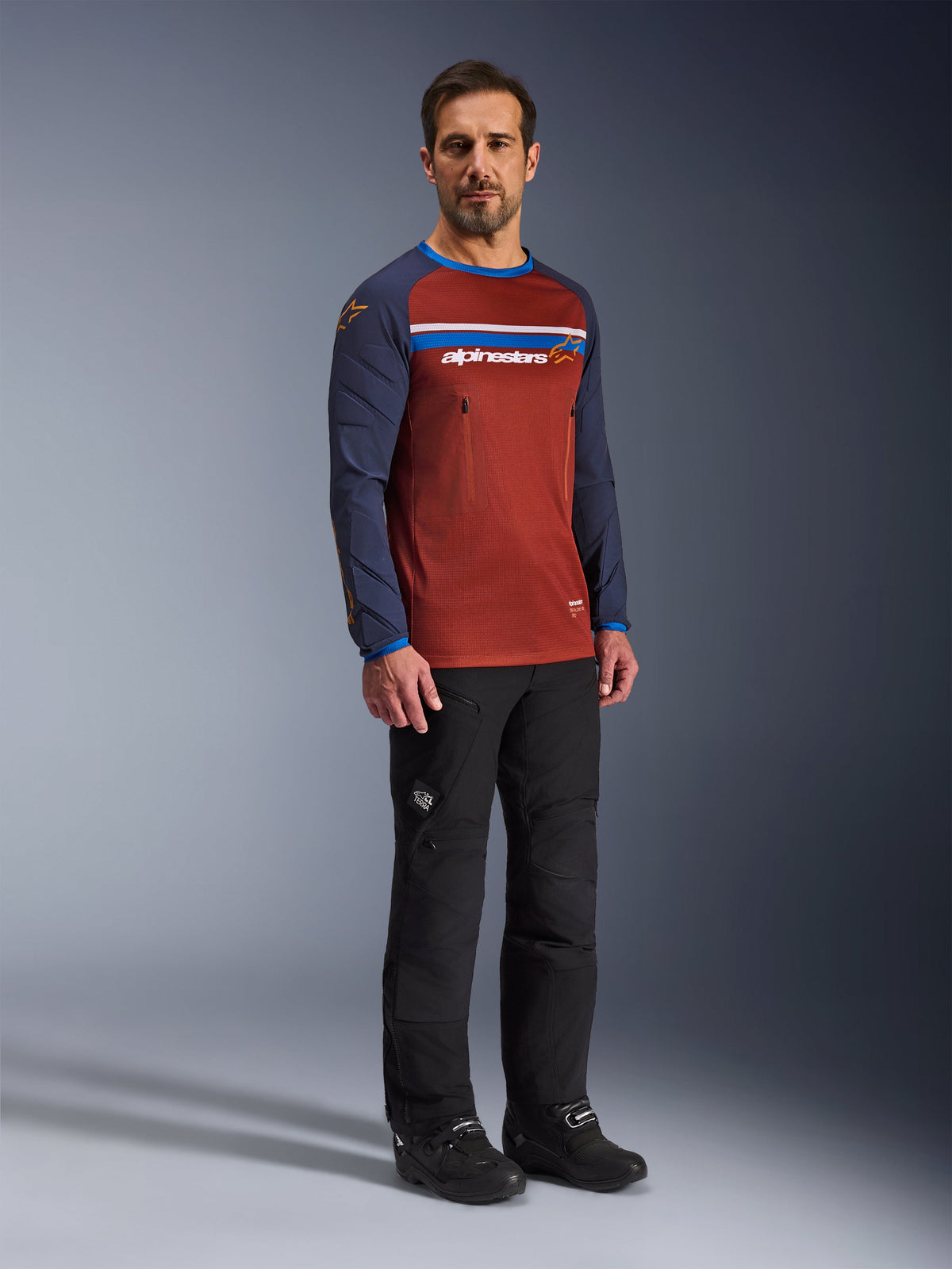 Mann in Alpinestars Maxdura Dual Jersey in Rust Brown und Mood Indigo, mit Mesh-Torso und gepolsterten Ärmeln, kombiniert mit schwarzer Motorradhose und Stiefeln, Adventure-Motorradbekleidung.
