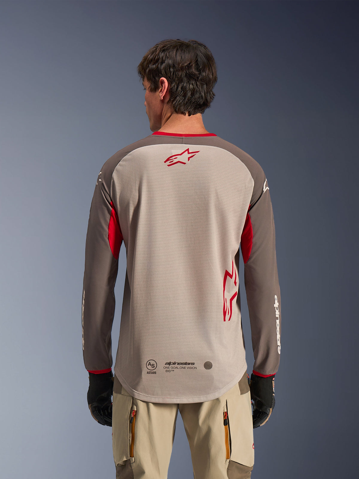 Rückansicht eines Mannes im Alpinestars Maxdura Dual Jersey in Aluminum Walnut, Adventure-Motorradtrikot mit hellbraunem Mesh-Torso und rotem Stern-Logo, dunkelbraune Ärmel mit roten Akzenten, beiger Fahrhose und schwarzen Handschuhen, entwickelt für erstklassige Offroad-Belüftung.