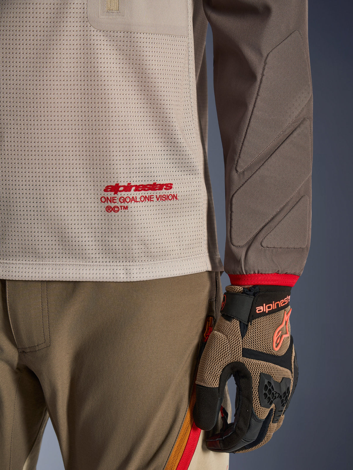 Alpinestars Maxdura Dual Jersey in Aluminum Walnut, Detailansicht des belüfteten Mesh-Torsos mit rotem Branding, abriebfester Ärmel mit integrierter Polsterung, getragen mit passender Adventure-Fahrhose und Handschuhen