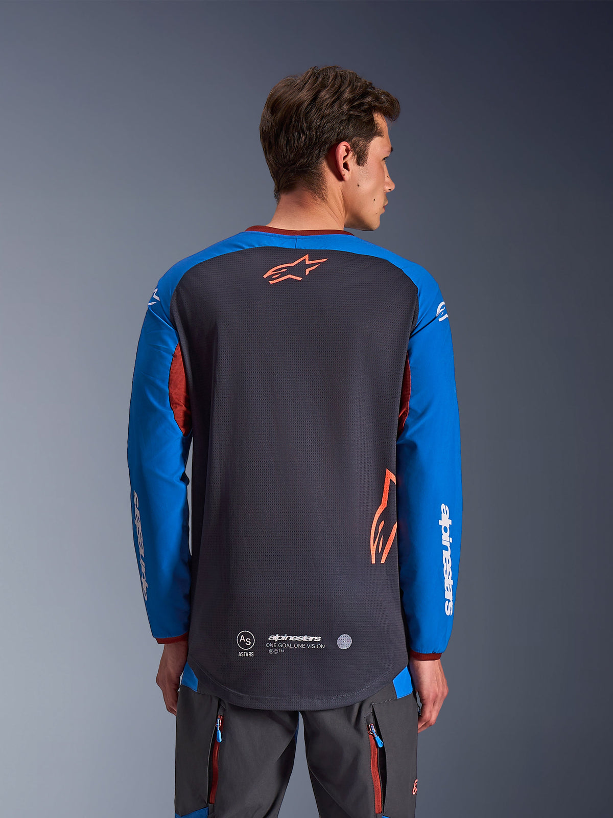 Alpinestars Maxdura Dual Jersey in Dunkelgrau und Royalblau, Rückansicht eines männlichen Modells, Mesh-Torso-Konstruktion mit orangefarbenem Astars-Logo, lange Ärmel mit weißem Branding, weinroter Kragen und Bündchen, getragen mit grauer technischer Hose, Dual-Sport- und Adventure-Motorradausrüstung