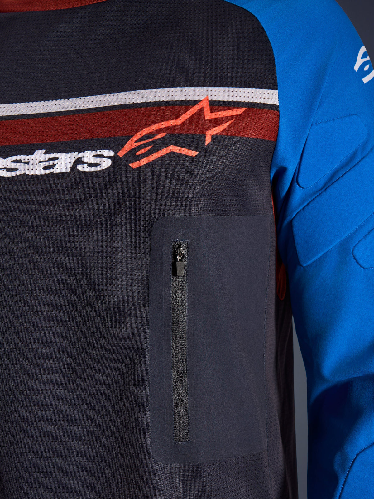 Alpinestars Maxdura Dual Jersey, Nahaufnahme von Brust und Ärmel, Dunkelgrau und Königsblau, mit Reißverschluss-Brusttasche, atmungsaktivem Mesh-Torso und integrierter Polsterung an den Ärmeln, Alpinestars-Logo auf roten und weißen Streifen.