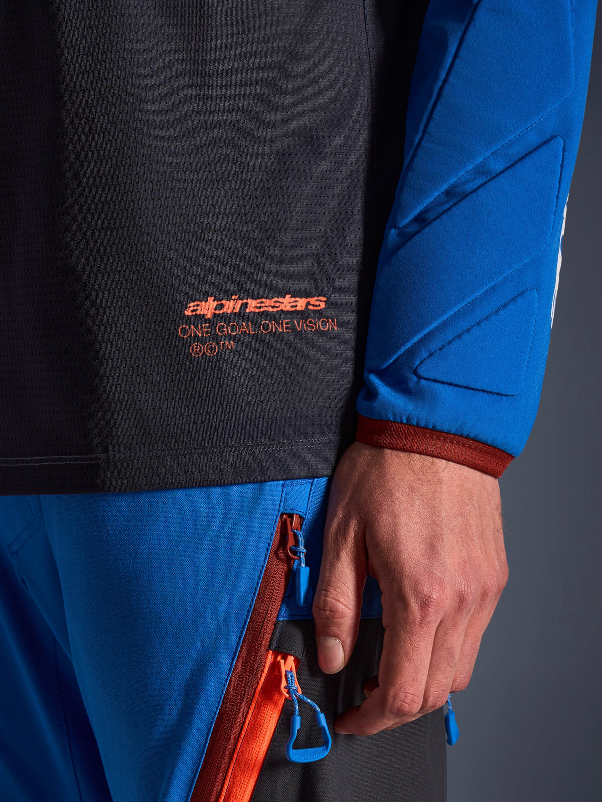 Nahaufnahme des Alpinestars Maxdura Dual Jersey in Deep Gray und Royal Blue, mit orangefarbenem Logo und 'One Goal One Vision' Schriftzug auf dem Mesh-Torso, gepolsterter Ärmel mit verstärkten geometrischen Einsätzen, technische Adventure-Fahrerausrüstung