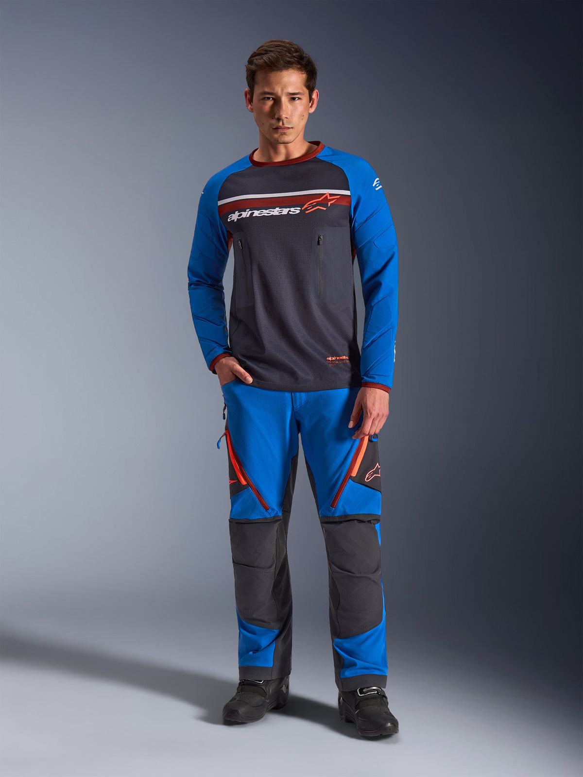 Alpinestars Maxdura Dual Jersey, Motorrad-Jersey, Dunkelgrau und Königsblau, Voll-Mesh-Torso mit gepolsterten Ärmeln und Brusttaschen mit Reißverschluss, weißes und orangefarbenes Alpinestars-Logo, getragen mit passender Adventure-Hose und schwarzen Stiefeln