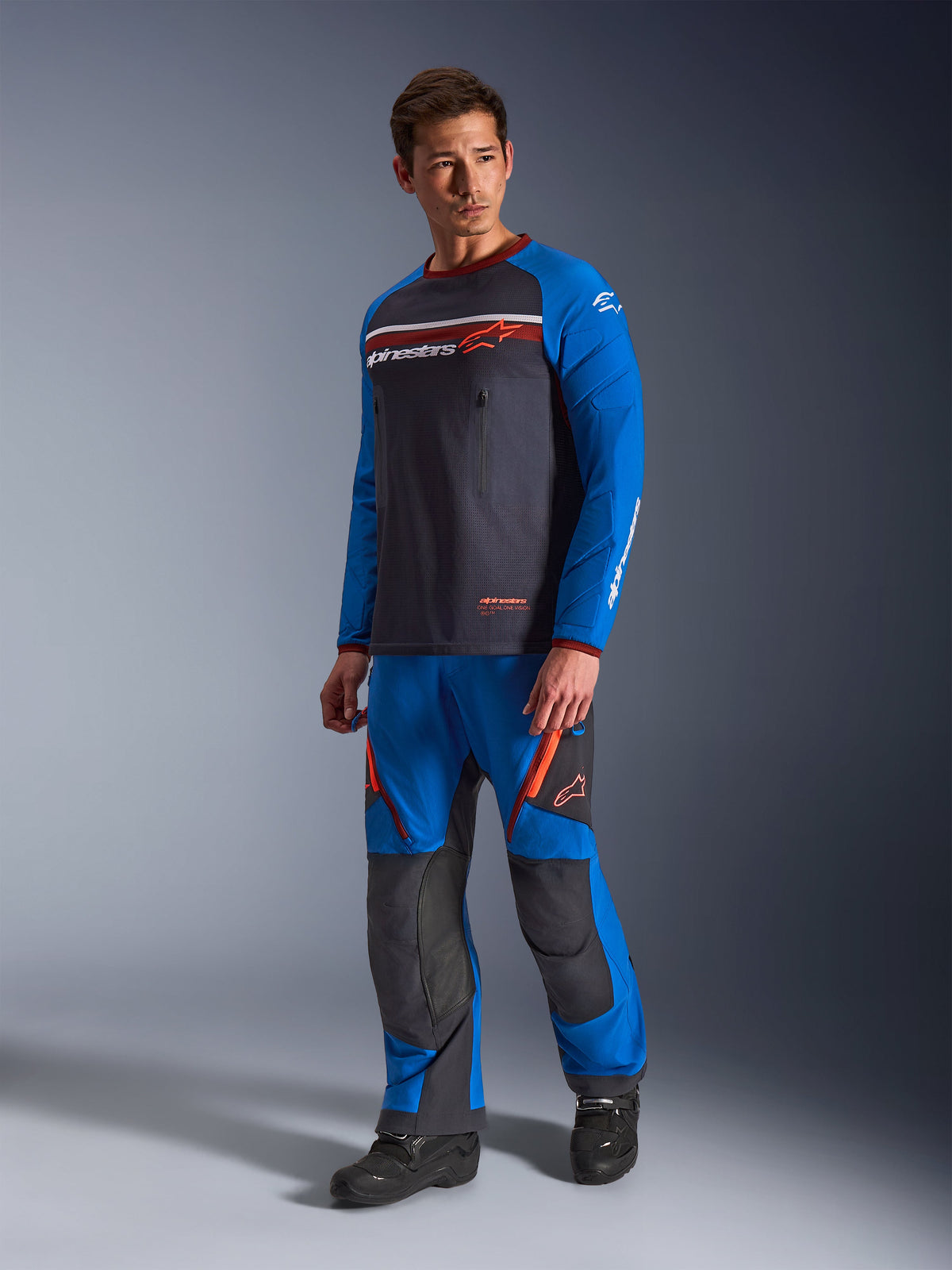 Alpinestars Maxdura Dual Jersey, Dunkelgrau und Royalblau, Adventure-Motorradtrikot mit Mesh-Torso und royalblauen Ärmeln, getragen von einem Model mit passender Offroad-Fahrhose, mit Brusttaschen mit Reißverschluss und Alpinestars-Branding.