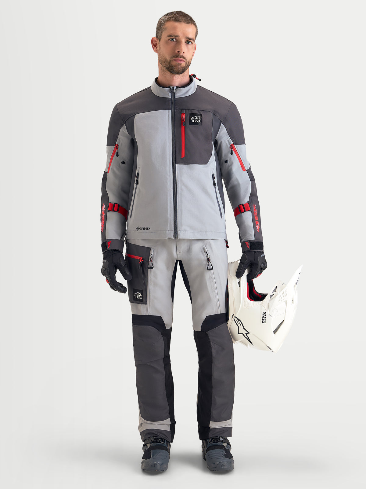 Alpinestars Arenis Gore-Tex Gear Set, men's adventure touring motorcycle jacket and pants in gray and dark gray with red accents, front full-length view on a model wearing matching riding gear, gloves, and holding a white helmet, waterproof all-terrain apparel.