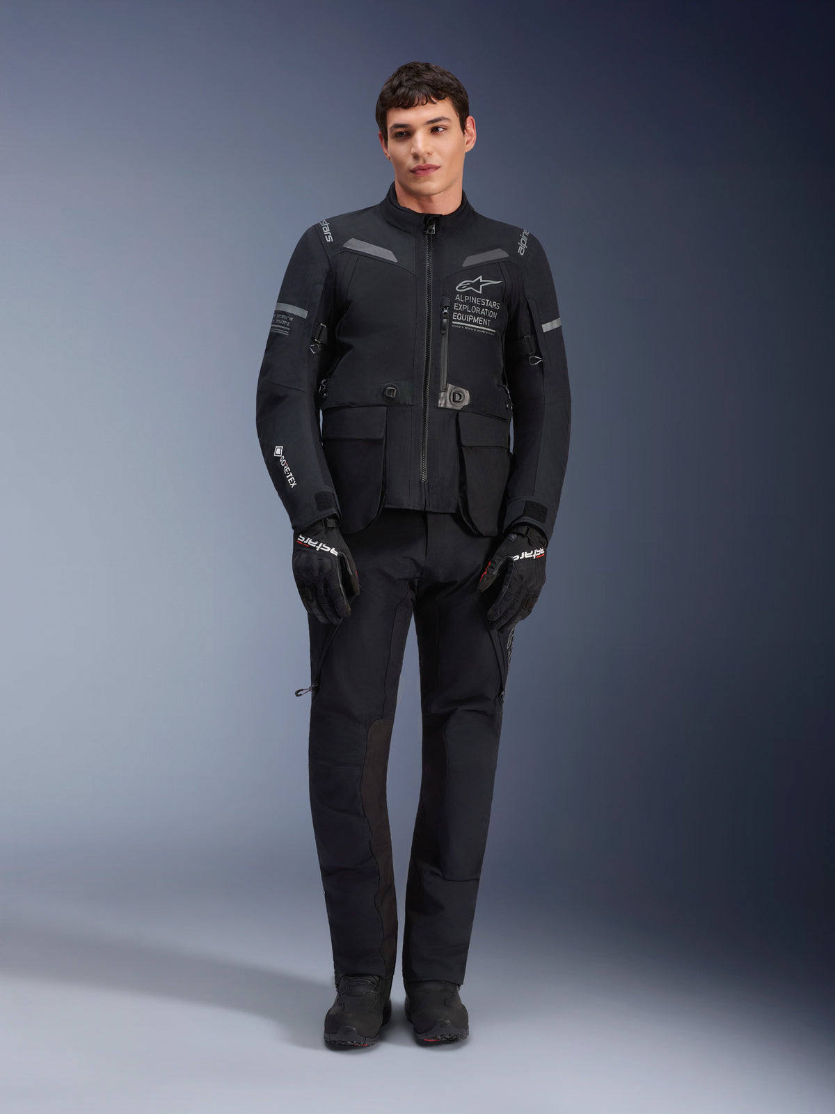 Alpinestars ST-7 2L GORE-TEX Jacket, adventure touring motorcycle jacket, Black and Dark Gray, worn with matching black riding pants and gloves, featuring multiple cargo pockets, reflective accents, and Alpinestars Exploration Equipment chest branding, functional design for road and off-road riding