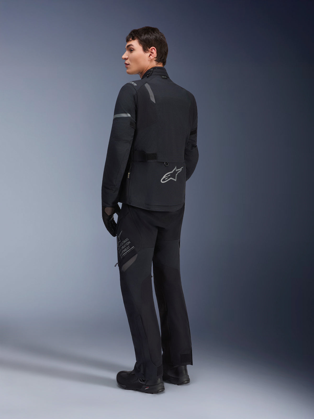 Male model wearing Alpinestars ST-7 2L GORE-TEX Jacket in Black Dark Gray, rear three-quarter view showing reflective accents and silver logo on lower back, adventure touring motorcycle apparel, paired with matching black riding pants and boots