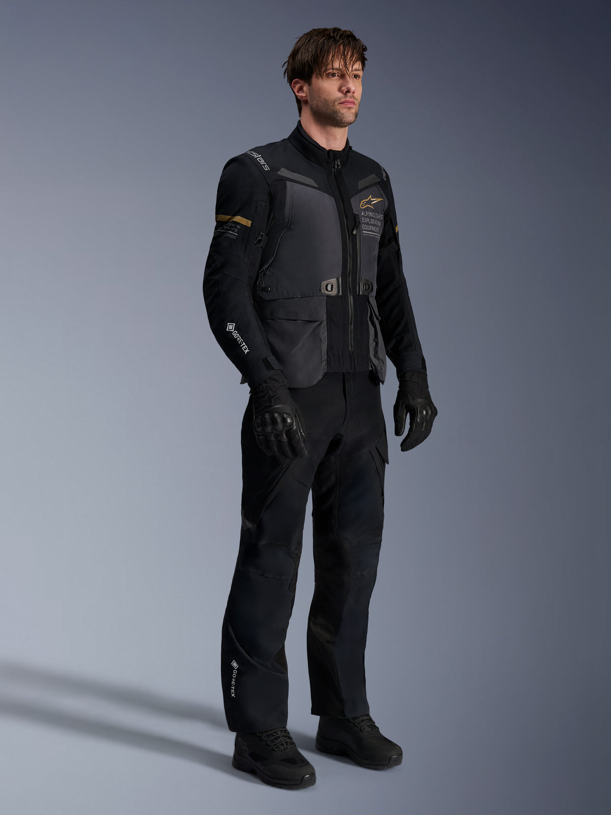 Männliches Model in der Alpinestars ST-7 2L GORE-TEX Jacke in Deep Gray Black und Dark Khaki, kombiniert mit passender schwarzer Adventure-Hose und Handschuhen, mit wasserdichter laminierter Außenschale, Cargotaschen und technischem Branding, ideal für Adventure-Touren.