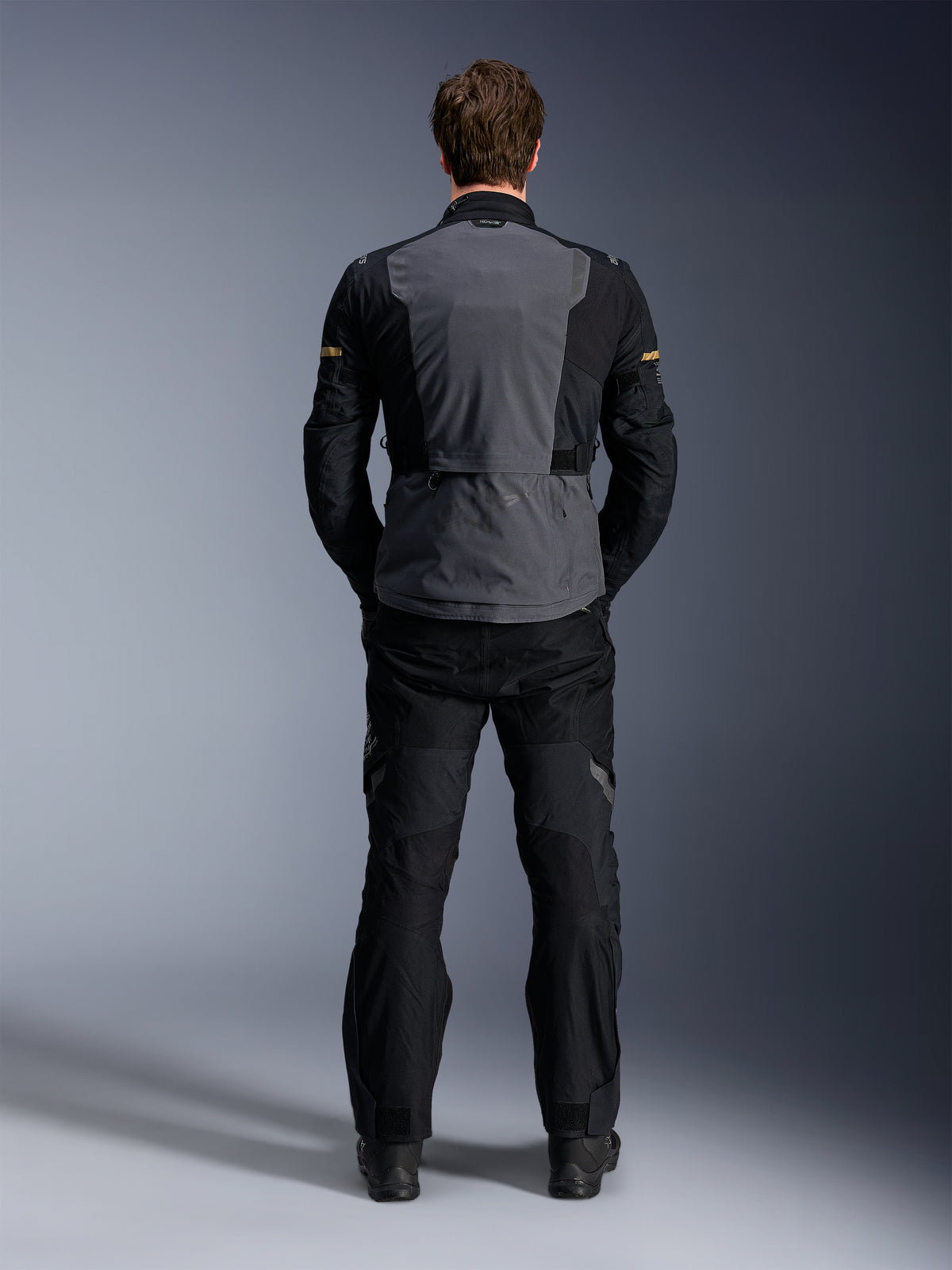 Alpinestars ST-7 2L GORE-TEX Jacke, Deep Gray Black und Dark Khaki, Rückansicht an einem Model, Abenteuer-Motorrad-Tourenjacke mit grauer Rückenpartie und schwarzen artikulierten Ärmeln, getragen mit passender schwarzer Hose.