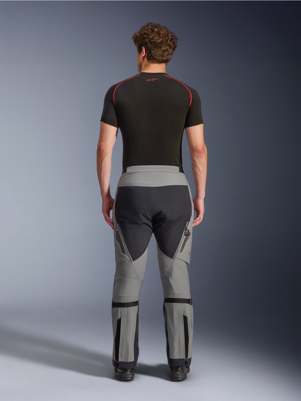 Alpinestars Nazca 3L GORE-TEX PRO Hose, Adventure-Motorradhose in Dunkelgrau und Schwarz, Rückansicht an einem Model, technisches Multi-Panel-Design mit grauem und schwarzem Gewebe, mit Wadenweitenverstellern und getragen mit einem schwarzen Baselayer mit roten Akzenten.