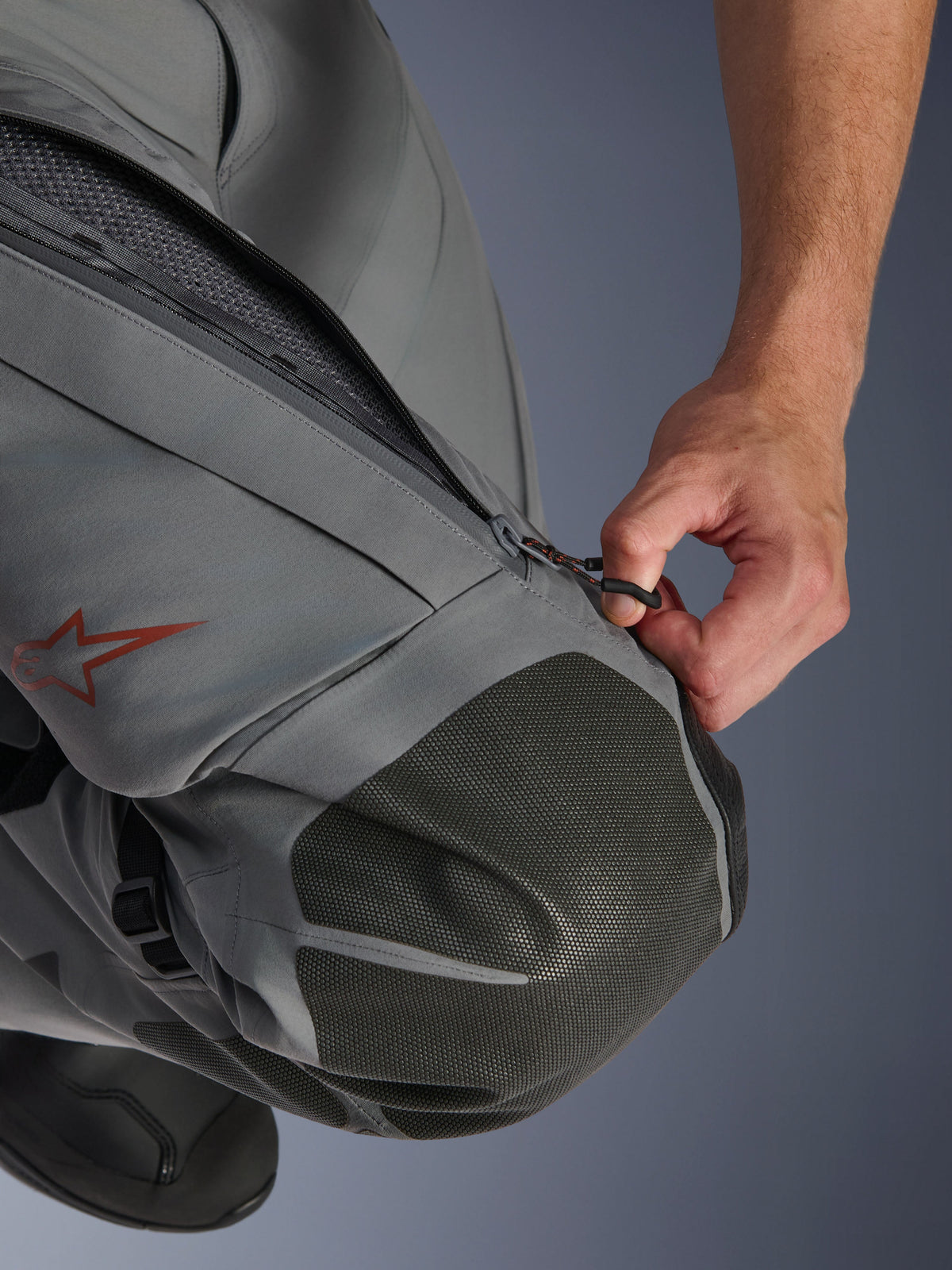 Alpinestars Nazca 3L GORE-TEX PRO Hose, Adventure-Motorradhose, Dunkelgrau und Schwarz, Nahaufnahme einer Hand, die den Belüftungseinlass mit Reißverschluss am Oberschenkel öffnet, mit strukturierten Superfabric-Knieverstärkungen und einem roten Alpinestars-Sternlogo, technische Textilkonstruktion für Allwetterschutz