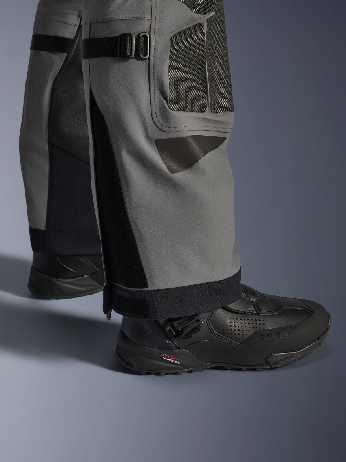 Alpinestars Nazca 3L GORE-TEX PRO Hose, Adventure-Motorradhose, Dunkelgrau und Schwarz, Nahaufnahme der Unterschenkel mit Wadenweitenverstellern und Superfabric®-Verstärkungen, getragen mit schwarzen Alpinestars-Stiefeln, strapazierfähiges Textil für extremes Adventure-Riding
