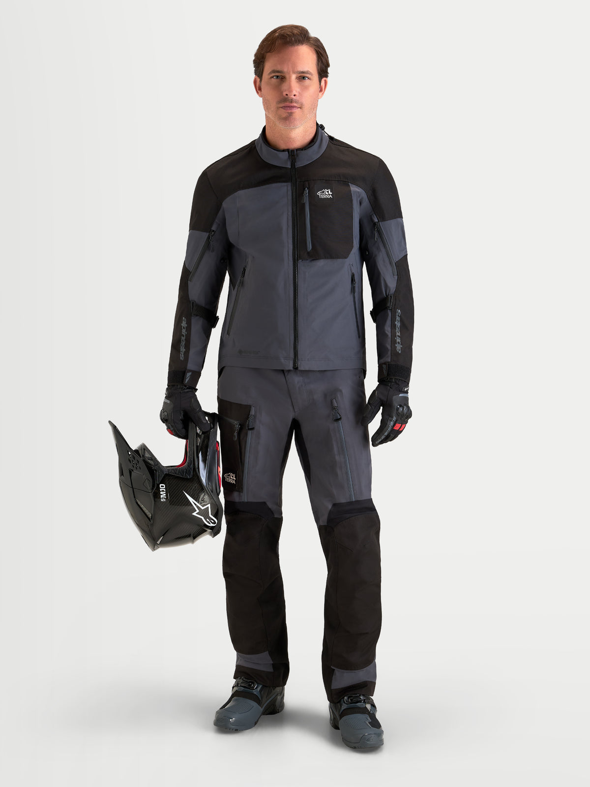 Alpinestars Arenis Gore Tex Hose, Herren-Adventure-Motorradhose in Schwarz und Dunkelgrau, Vorderansicht an einem Model mit passender Ausrüstung, Helm und wasserdichter Textilbekleidung