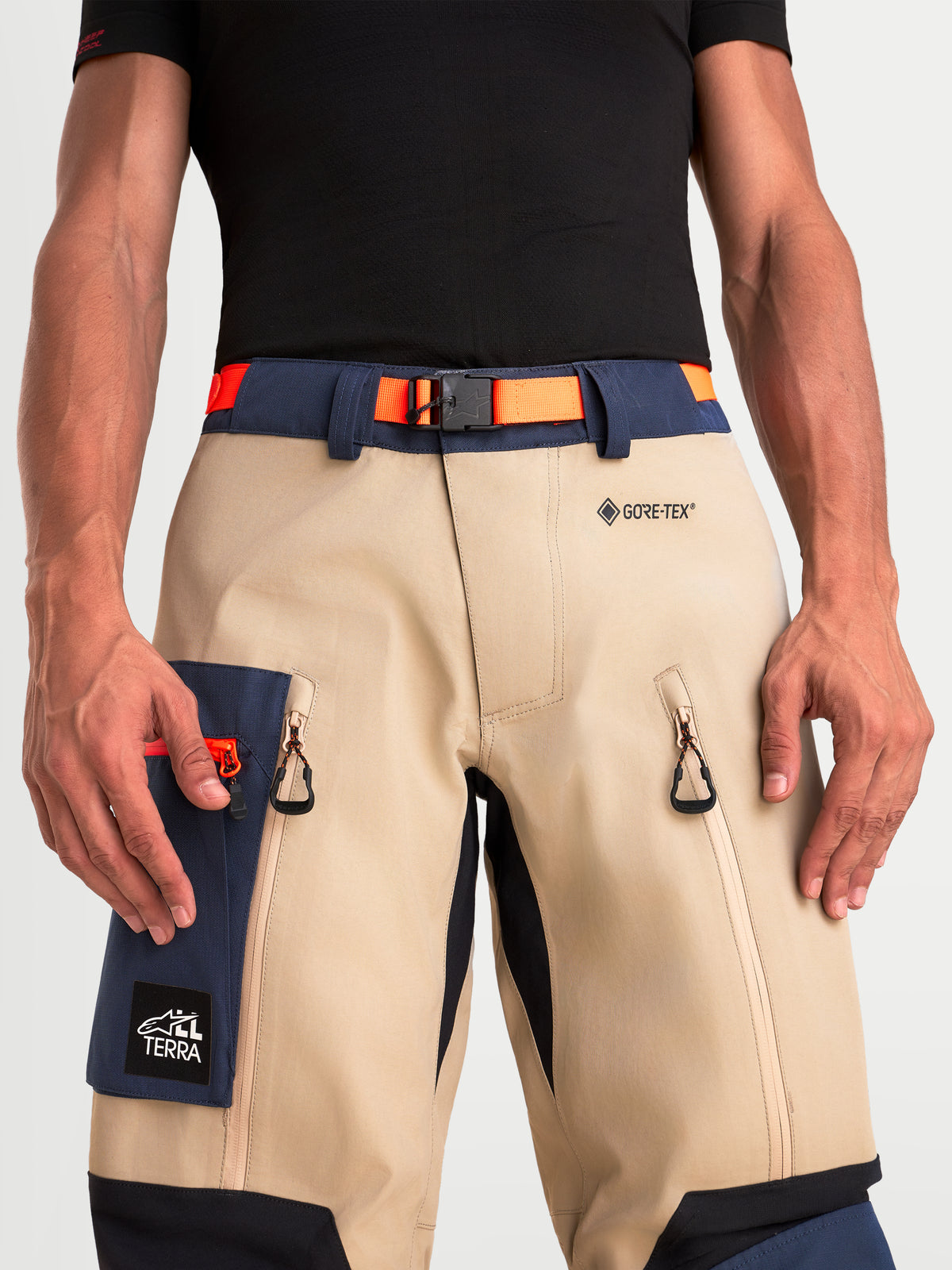 Alpinestars Arenis Gore-Tex Hose, Herren-Adventure-Motorradhose in Beige und Marineblau mit orangefarbenem Taillengürtel, Nahaufnahme mit GORE-TEX-Branding, Magnetverschluss, Oberschenkelbelüftungen und Cargotasche an Model