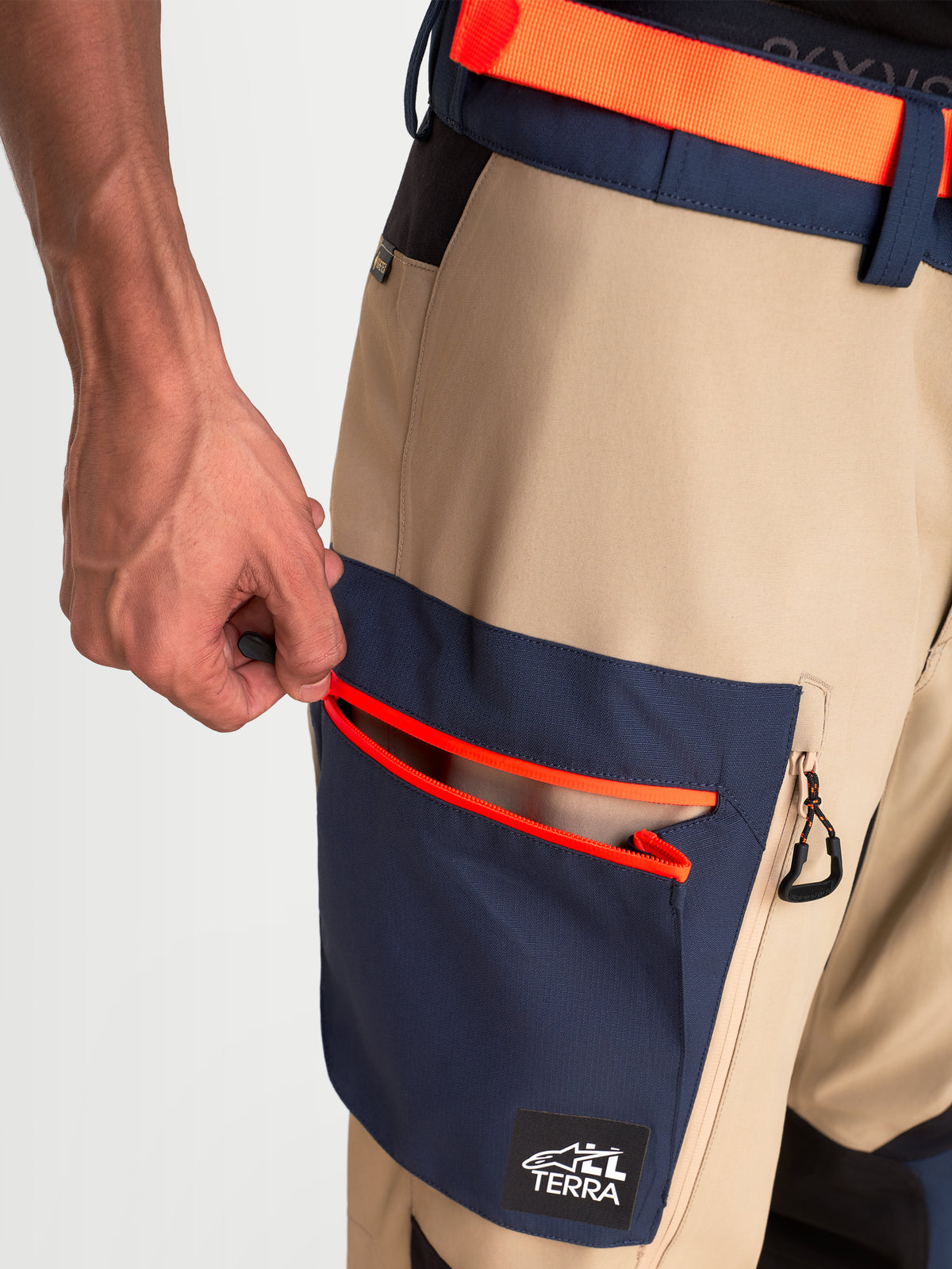 Alpinestars Arenis Gore-Tex Hose, Adventure-Touren-Motorradhose, Nahaufnahme zeigt eine Hand, die die integrierte Cargotasche mit leuchtend orangen Reißverschlussdetails öffnet, in Beige und Marineblau