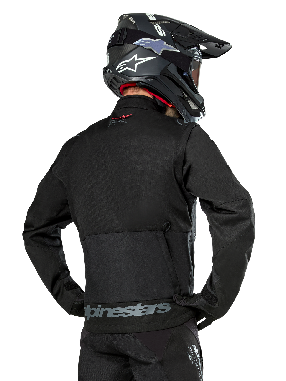 Back view of a rider wearing the Alpinestars Pro-Dura Jacket in Black, durable ripstop textile enduro jacket, featuring a large rear cargo pocket with gray Alpinestars branding across the lower back, paired with a black motocross helmet.