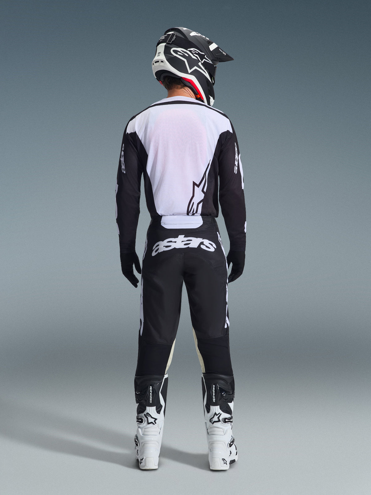 Motocross-Fahrer in der Rückansicht mit Alpinestars 2026 Racer Air Riway Hose in Schwarz und Weiß, passendem schwarz-weißem Langarm-Trikot, weiß-schwarzen Rennstiefeln und schwarzem Helm mit Alpinestars-Branding.