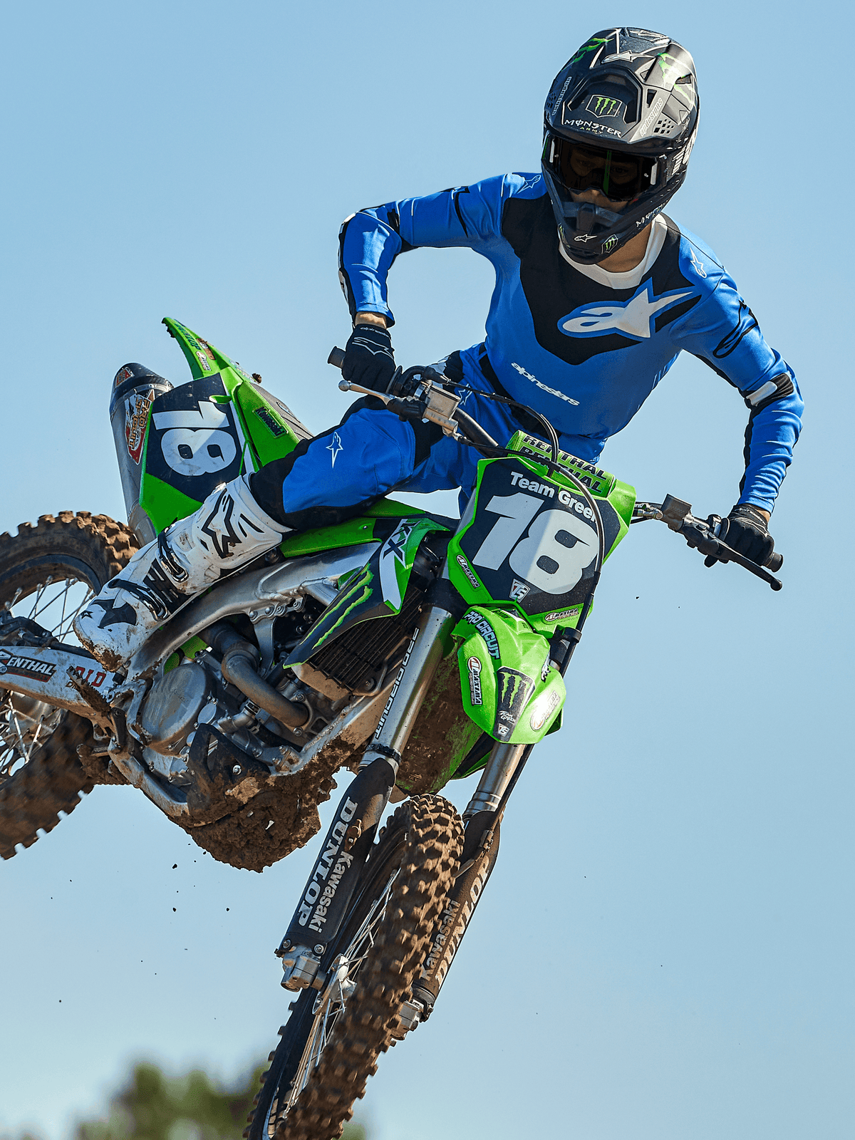 Motocross-Fahrer mitten im Sprung auf einem grünen Motorrad, trägt Alpinestars 2025 Racer Veil Hose in Blau und Schwarz, passendes blaues Trikot mit weißem Brustlogo, weiß-schwarze Stiefel und schwarzen Helm, Hochleistungs-Offroad-Ausrüstung für den Rennsport.