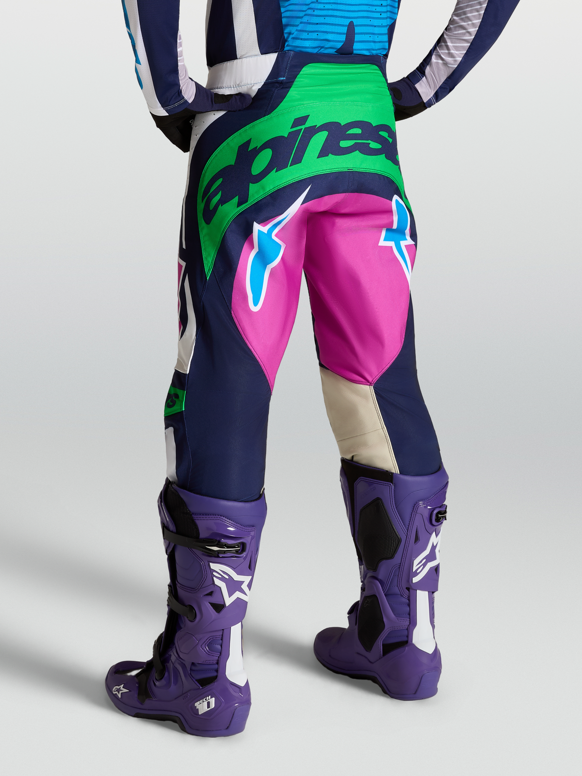 Alpinestars Techstar Maven Hose, Motocross-Ausrüstung in Blau, Grün und Pink, Rückansicht am Model mit violetten Tech 10 Stiefeln, strapazierfähige Offroad-Bekleidung.