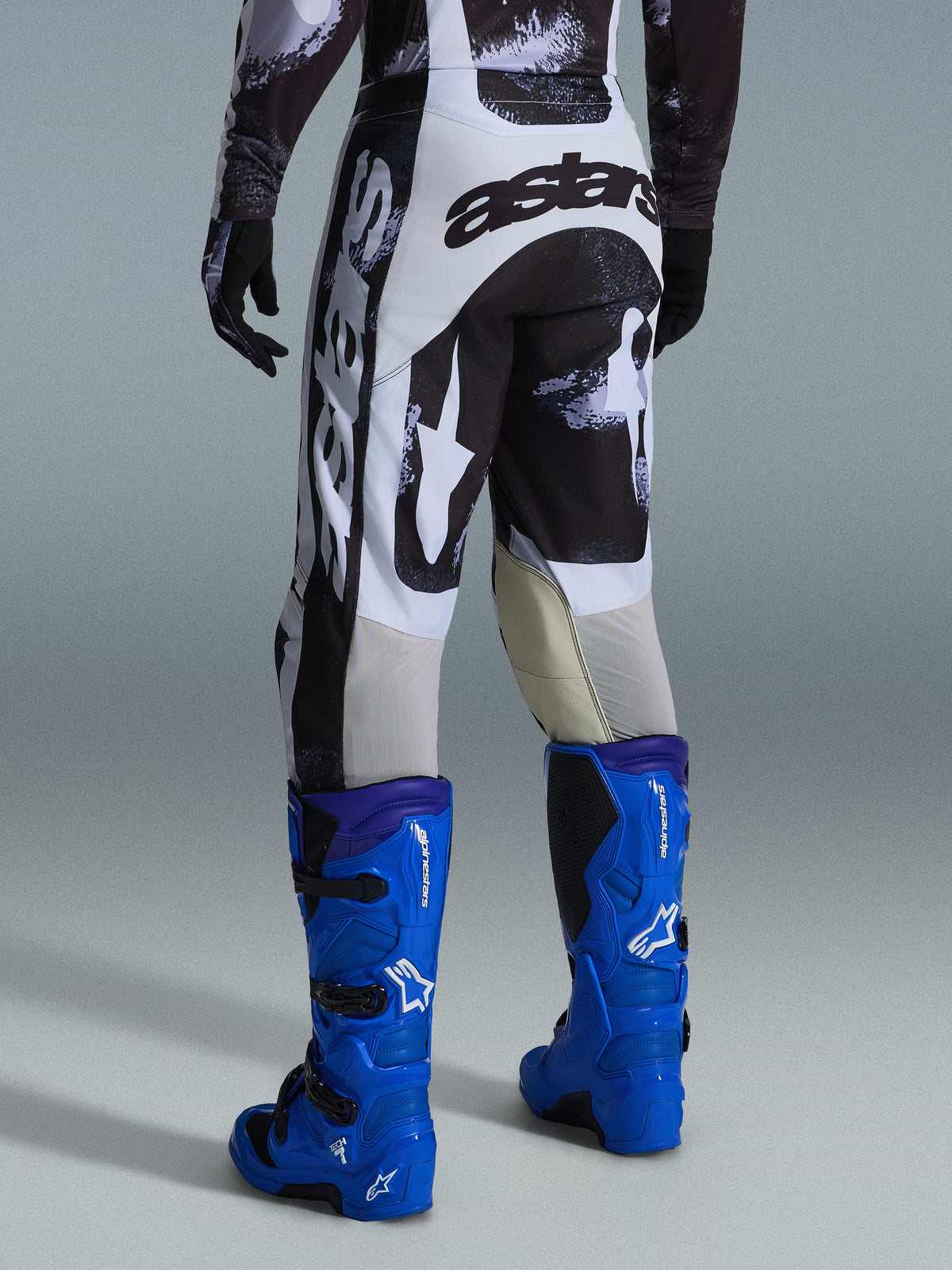 Alpinestars 2026 Racer Lahnd Hose, Motocross-Hose, Iron Camo Muster in Grau und Schwarz, Rückansicht mit großem schwarz-weißem Astars-Branding, kombiniert mit leuchtend blauen Motocross-Stiefeln, professionelle Offroad-Rennausrüstung