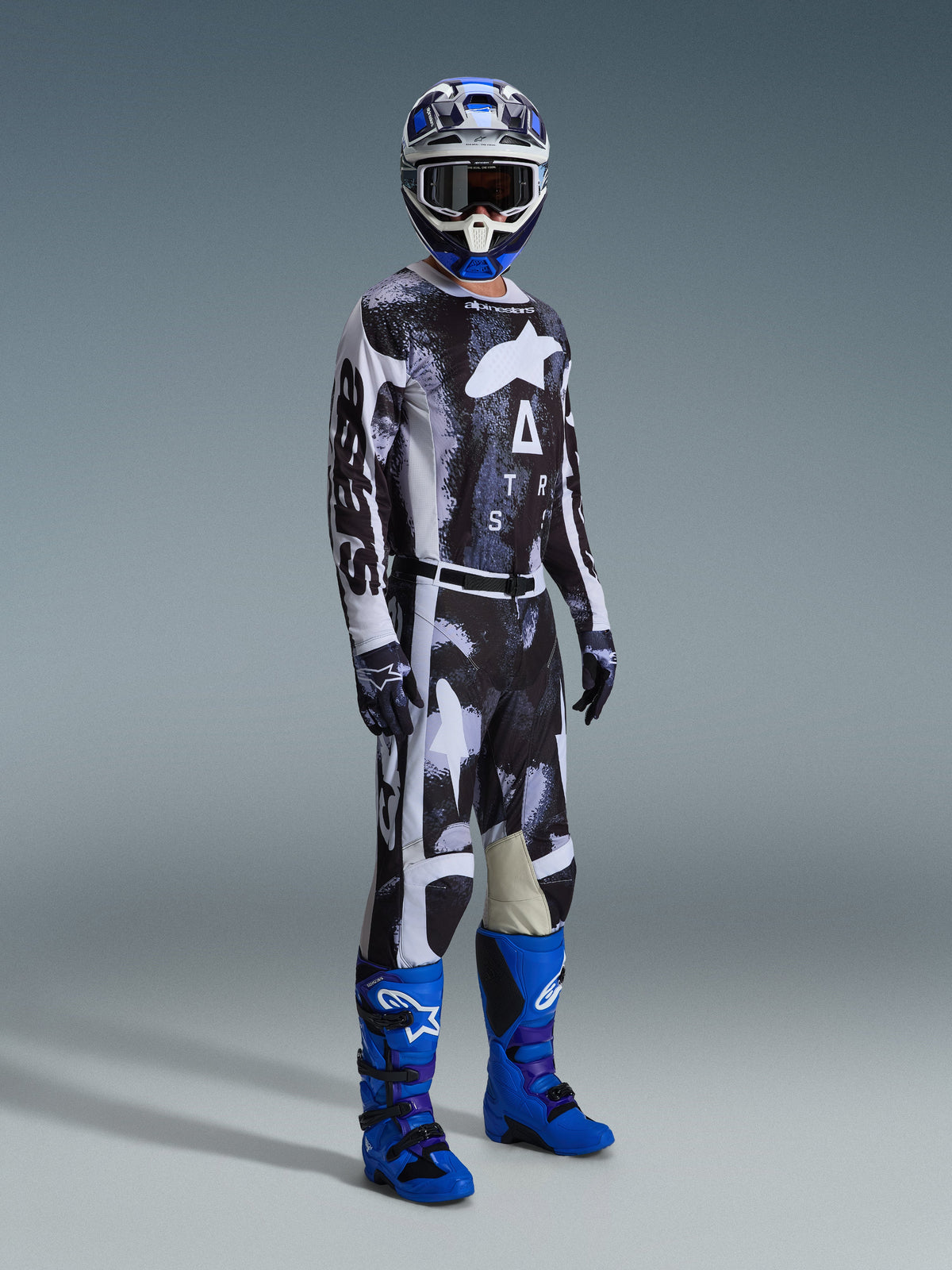 Motocross-Fahrer im Alpinestars 2026 Racer Lahnd Trikot und passender Hose in Iron Camo mit schwarz-weißem Grafikmuster, kombiniert mit blauen Stiefeln, einem blau-weißen Helm und schwarzen Handschuhen, komplette Rennausrüstung für den Offroad-Einsatz.