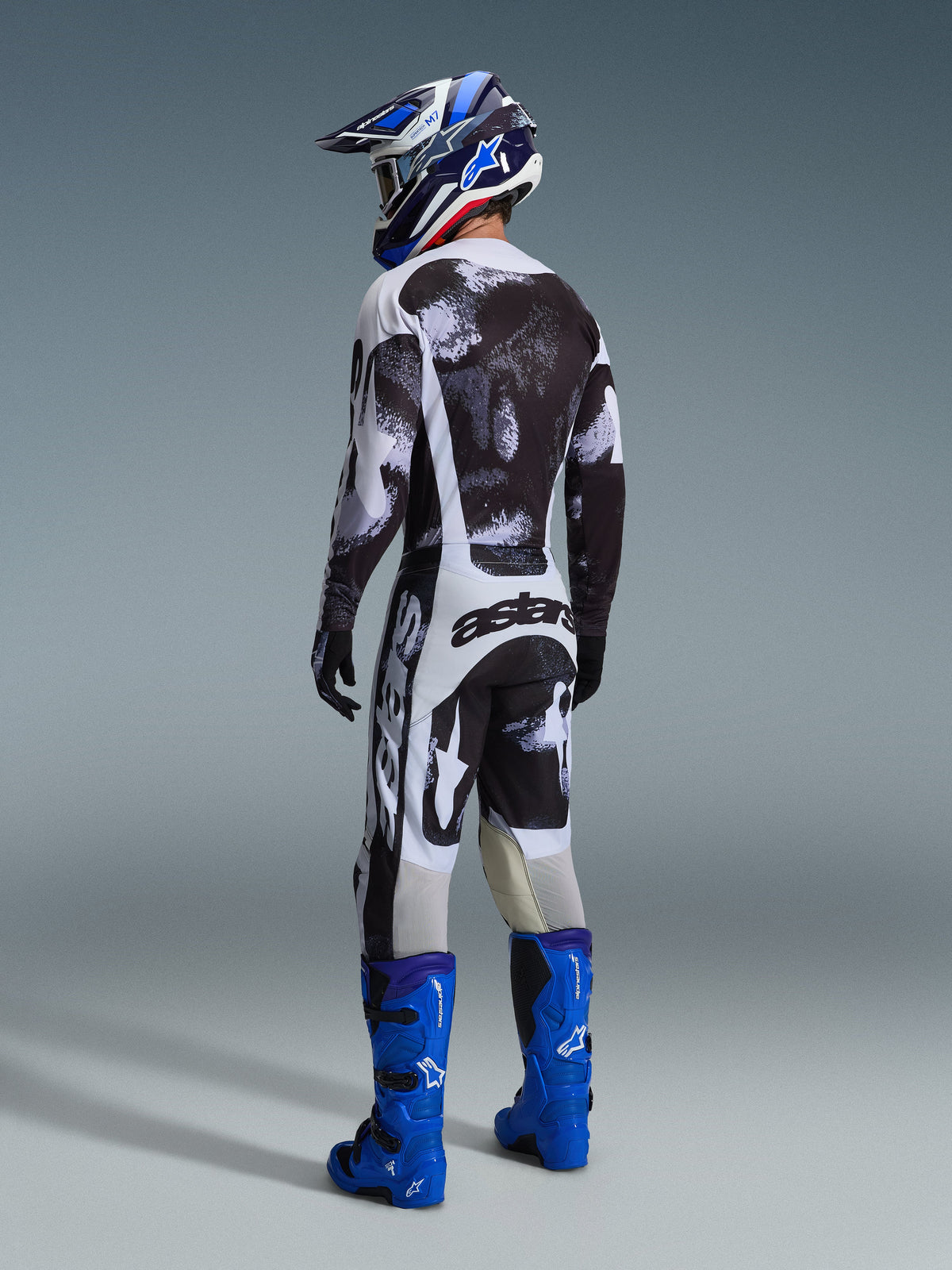 Motocross-Fahrer in der Rückansicht mit Alpinestars 2026 Racer Lahnd Hose in Iron Camo, passendem schwarz-weiß gemustertem Trikot, leuchtend blauen Tech-Stiefeln und blau-weißem Helm, abgestimmte Offroad-Rennausrüstung mit markantem „astars“-Branding am Gesäß.