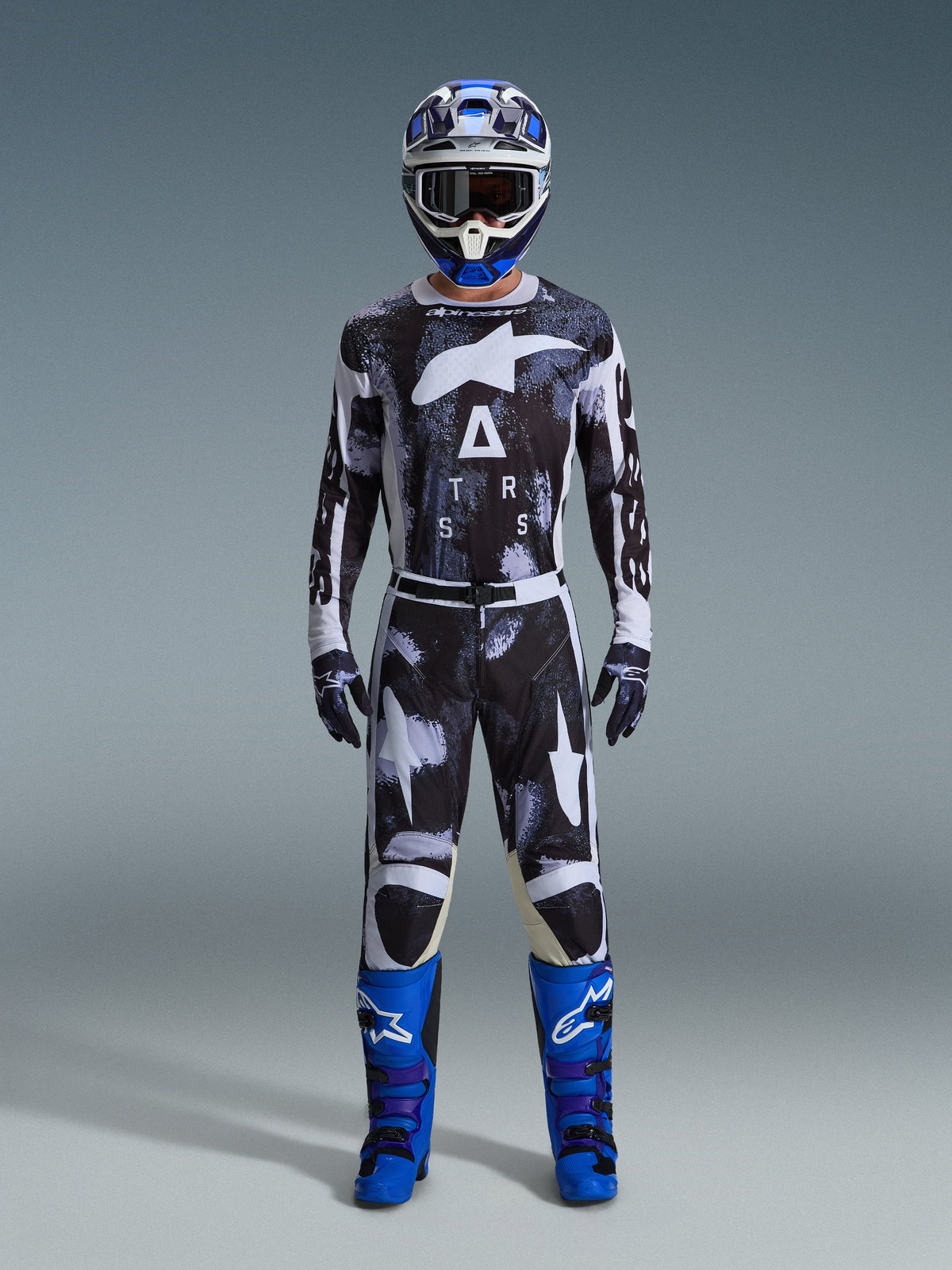 Motocross-Fahrer mit Alpinestars 2026 Racer Lahnd Hose in Iron Camo, grau-schwarzes Camouflage-Design, passendes Trikot, blau-weißer Helm mit Schutzbrille und leuchtend blaue Stiefel, abgestimmtes Motocross-Ausrüstungsset.