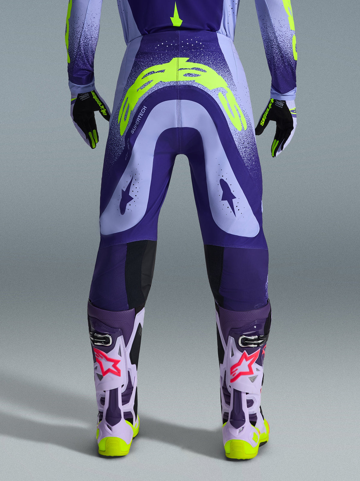 Alpinestars 2026 Supertech Scenz Hose, Motocross-Hose, Lila und Gelb Fluo, Rückansicht mit großem gelben Logo am Gesäß, getragen mit passendem Trikot, Handschuhen und Schutzstiefeln, athletische Passform für professionellen Motocross-Rennsport.