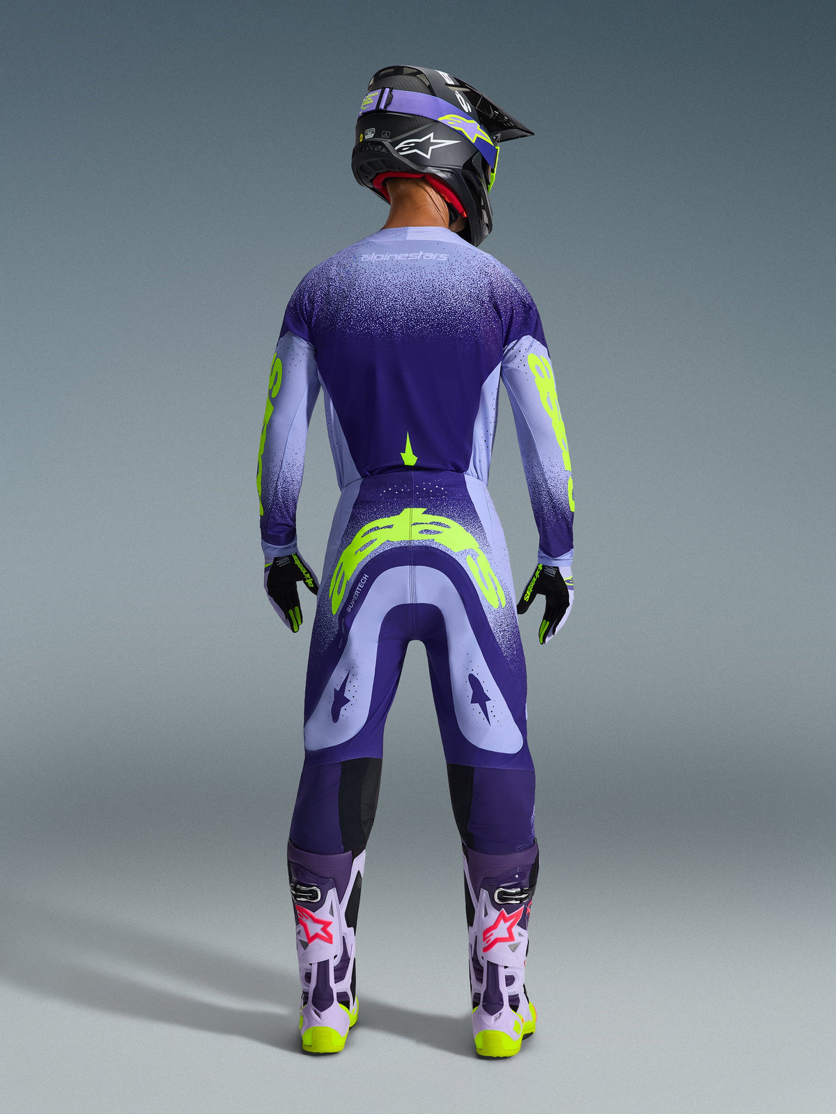 Motocross-Fahrer von hinten in Alpinestars 2026 Supertech Scenz Hose in Lila Gelb Fluo, passendes Trikot mit lila und neongelben Grafiken mit Farbverlauf, schwarzer Helm und lila Stiefel, professionelle Rennausrüstung für Offroad-Performance