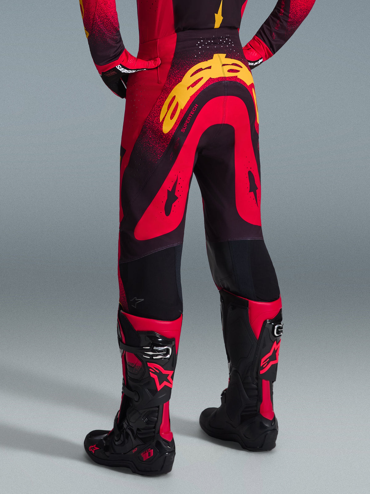 Alpinestars 2026 Supertech Scenz Hose, Motocross-Hose, Rot, Schwarz und Gelb, Rückansicht mit großem gelben Alpinestars-Branding auf der Gesäßpartie, kombiniert mit passendem Jersey und rot-schwarzen Motocross-Stiefeln.